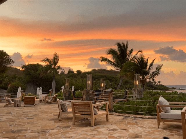 A sandy beach with various lounge chairs, palm trees and lanterns in the distance. There is also a gorgeous orange sunset.