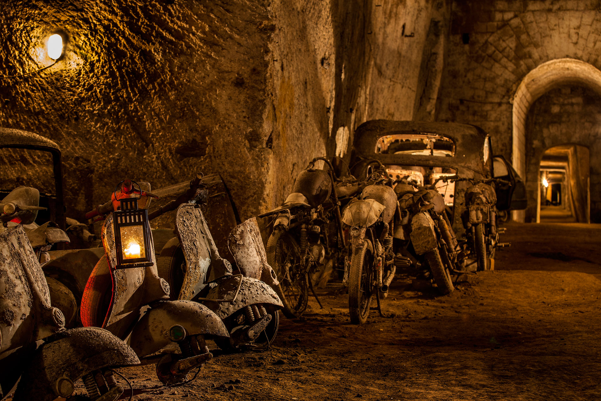 A view of a rocky tunnel with old vespas, lanterns and cars leading towards an archway.