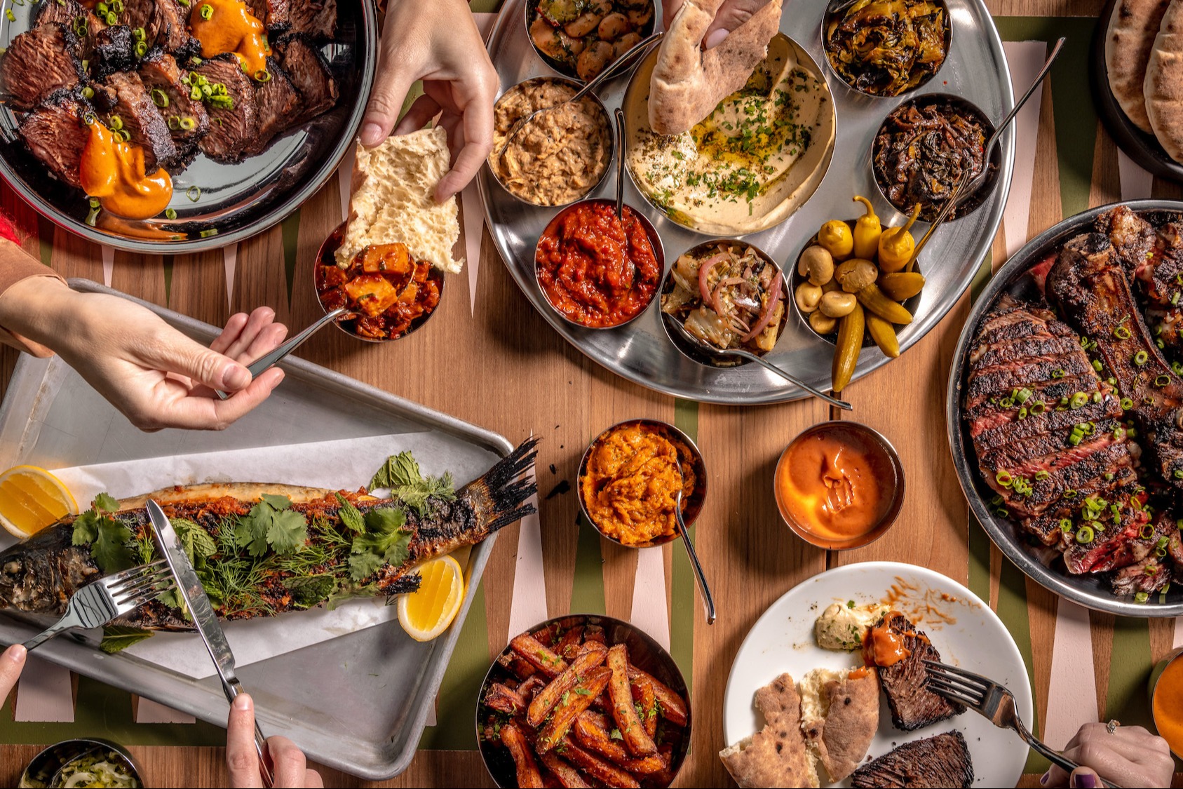 A wooden table full of various platters of food and hands cutting and scooping items with dining utensils.