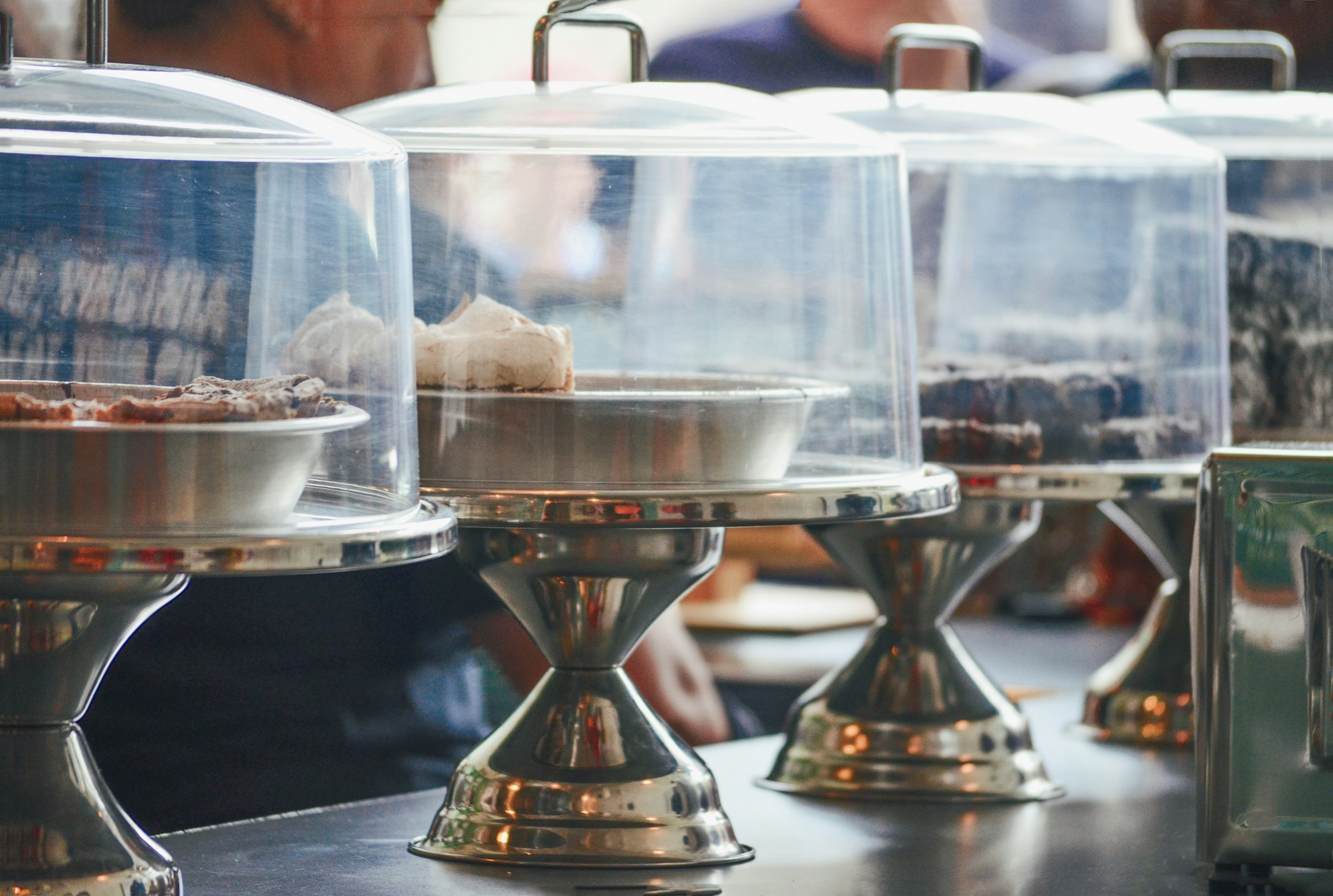 A row of cake stands full of various pies in platters covered by a clear lid.