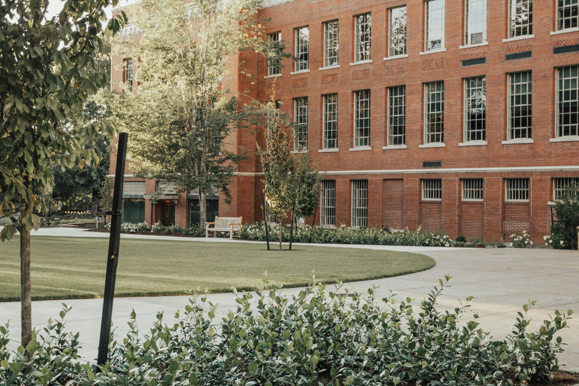A red brick building with various windows. There is also a curved path, manicured lawn, trees, bushes and wooden bench in the surrounding areas.