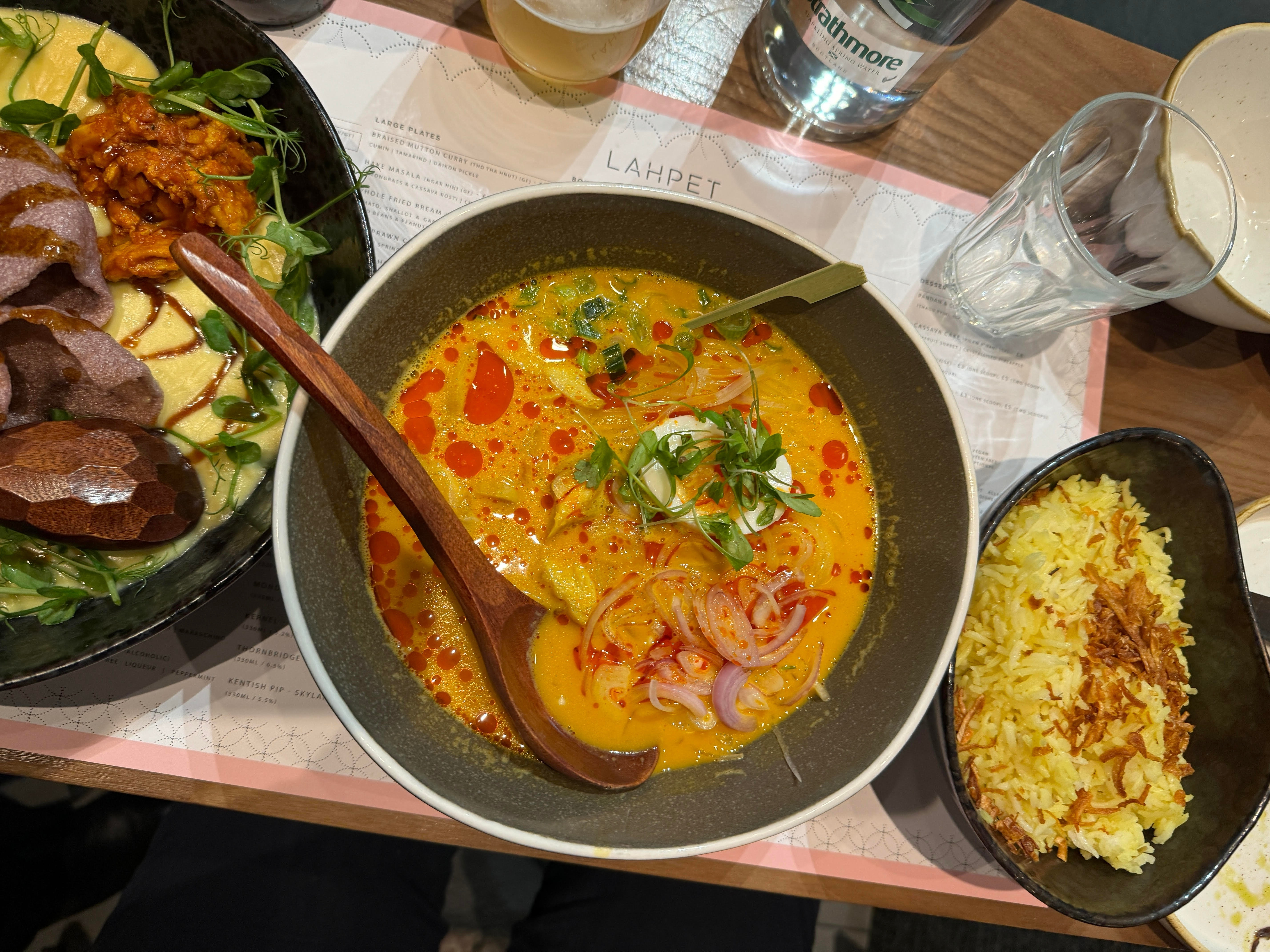 A picture of a table complete with glassware, a menu and three bowls of food that appear to be rice, curry and a meat dish with wooden utensils.