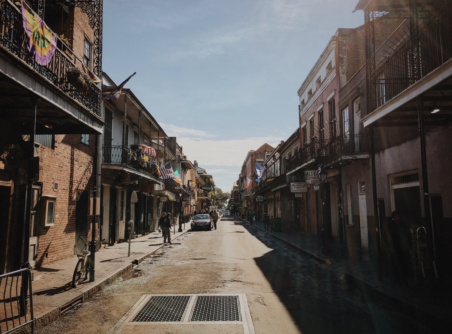 A rustic view of an old street surrounded by brick buildings with metal railing, hanging plants and parked cars with people walking around.