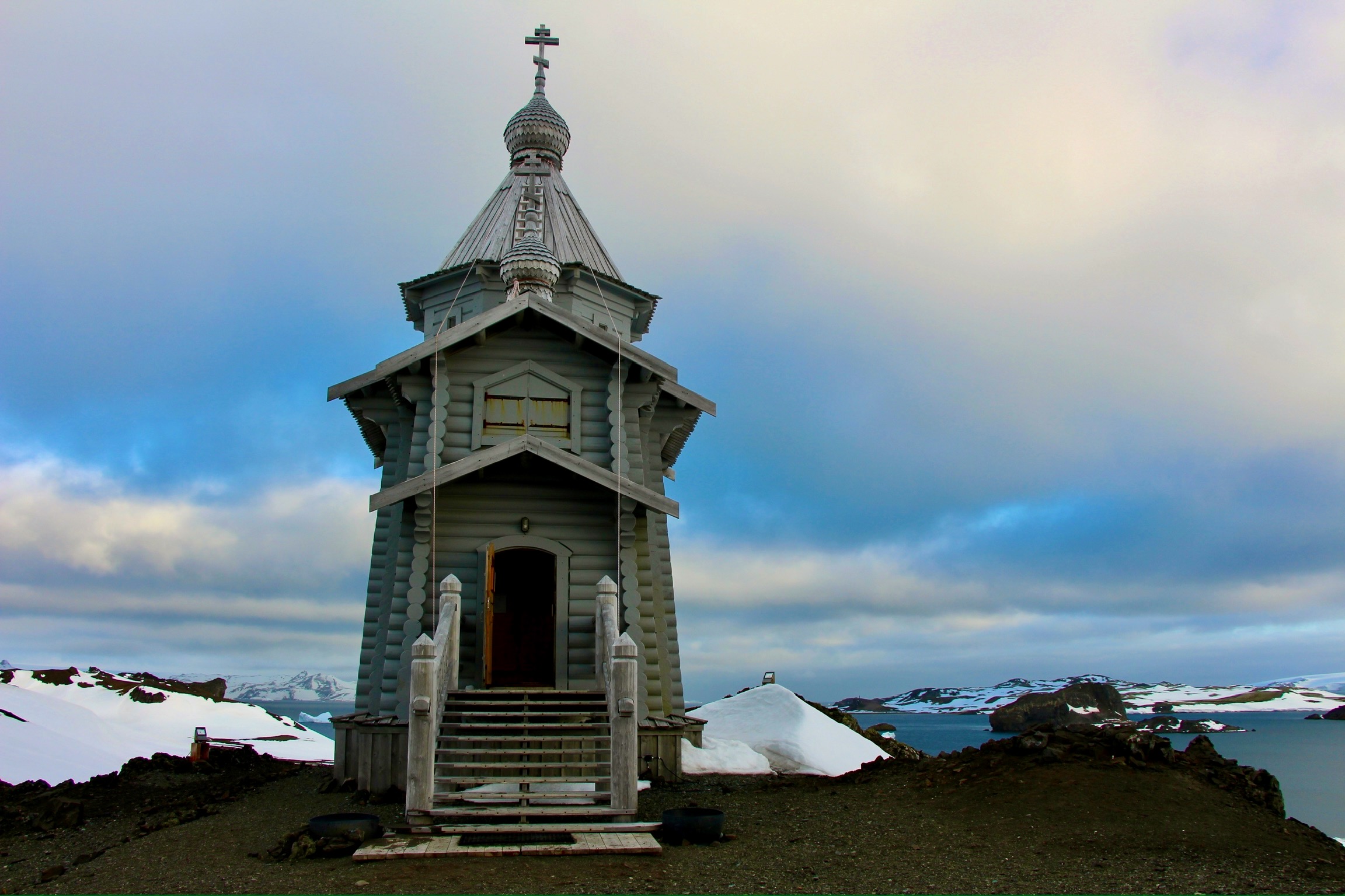 A view of a small, green church on top of a dirt ground with snow, water and ice surrounding it.