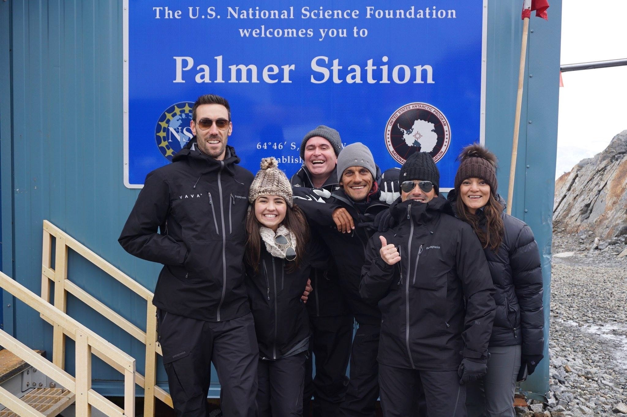 A picture of a group of six people standing in front of a building and blue sign that reads Palmer Station.
