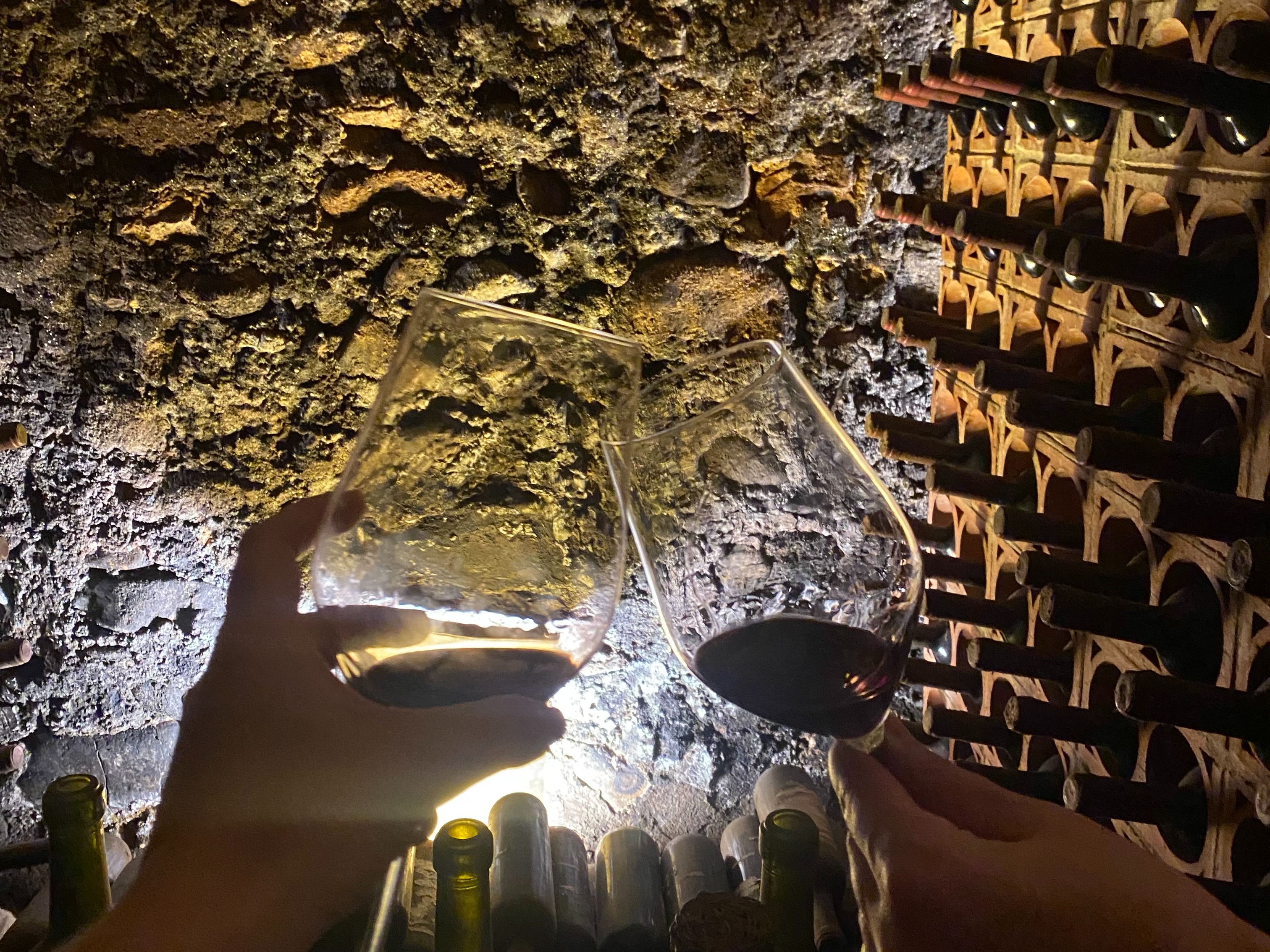 Two people cheersing with their wine glasses inside of a cave that is also a wine cellar.