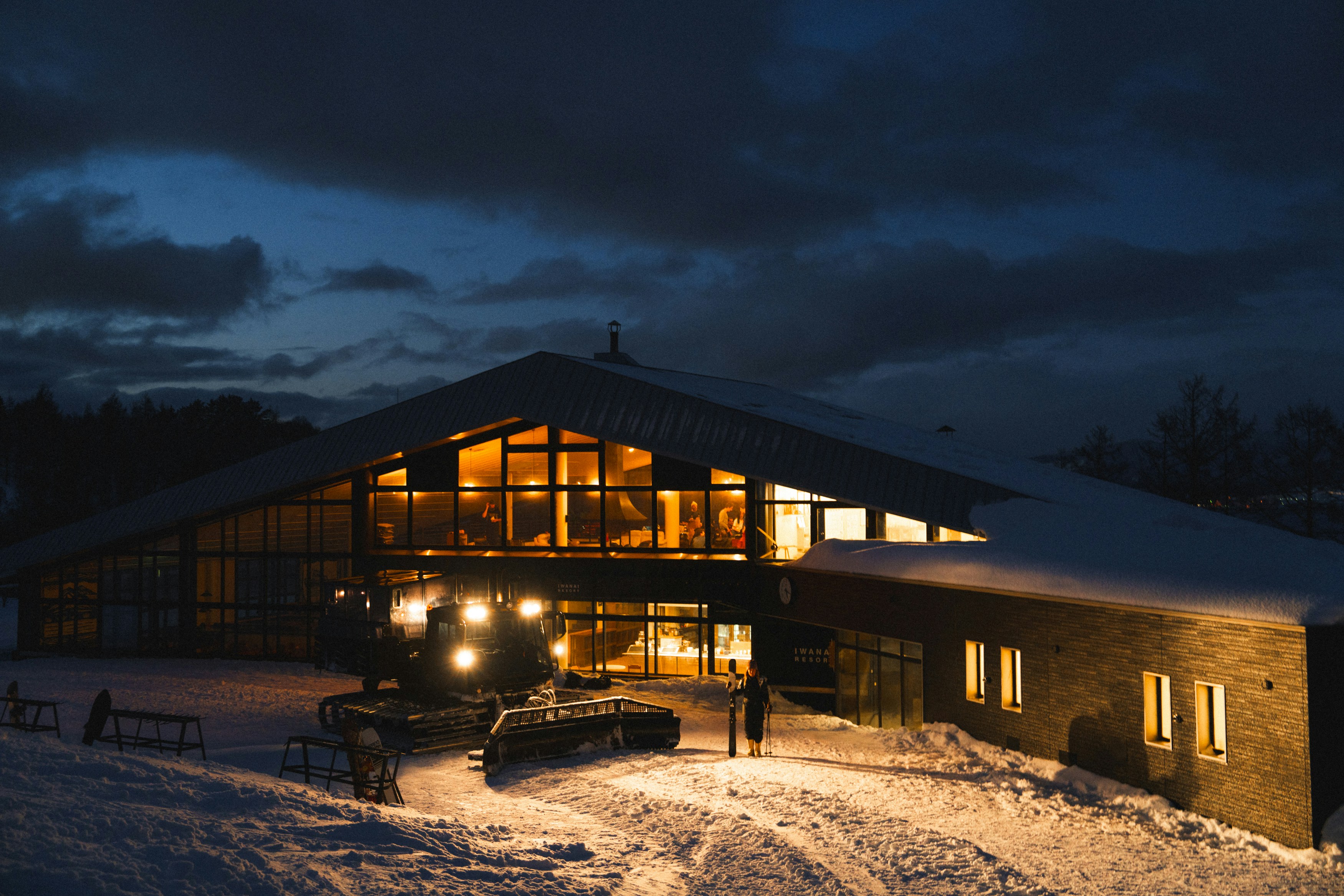 A view of a ski lodge at nighttime with large windows lit up from the inside and a snowy driveway in the foreground.