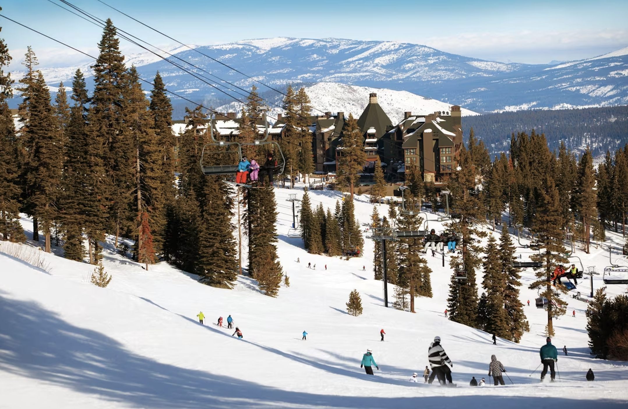 A beautiful ski hill surrounded by lush, green pine trees and various people in colorful ski gear on the snowy hill. There is also a snowy covered mountain in the far distance.