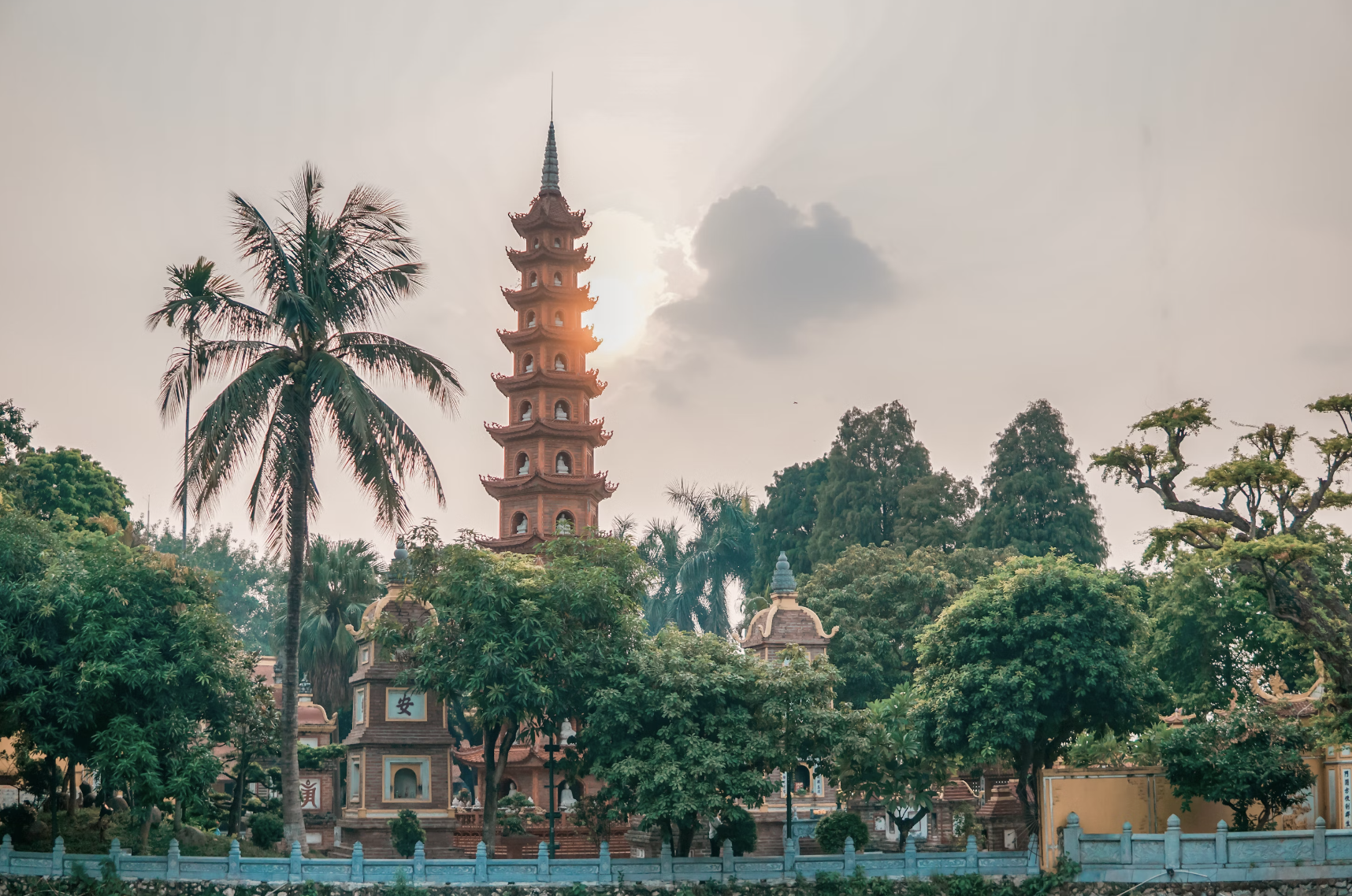 A beautiful sunset view of a large temple surrounded by lush trees, in particular one large palm tree and a metal fence.