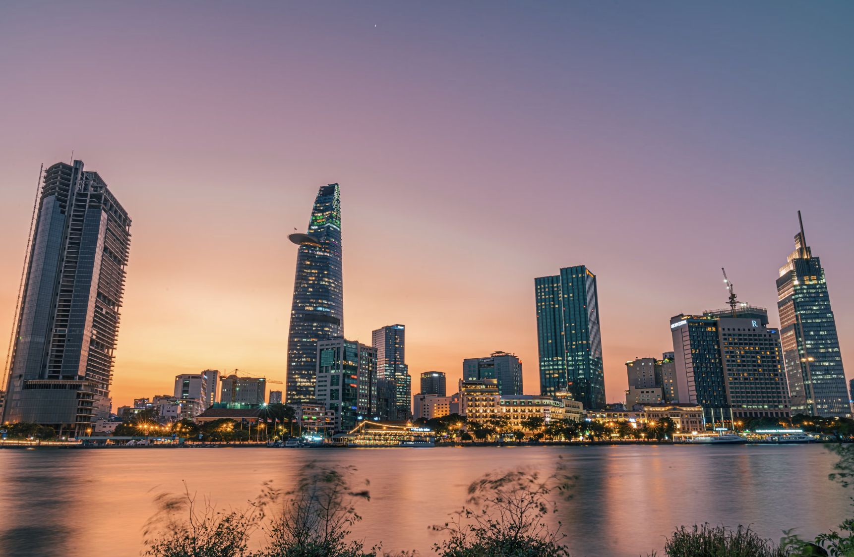 A sunset view of a city skyline in Ho Chi Minh with a still body of water and bushes in the forefront.