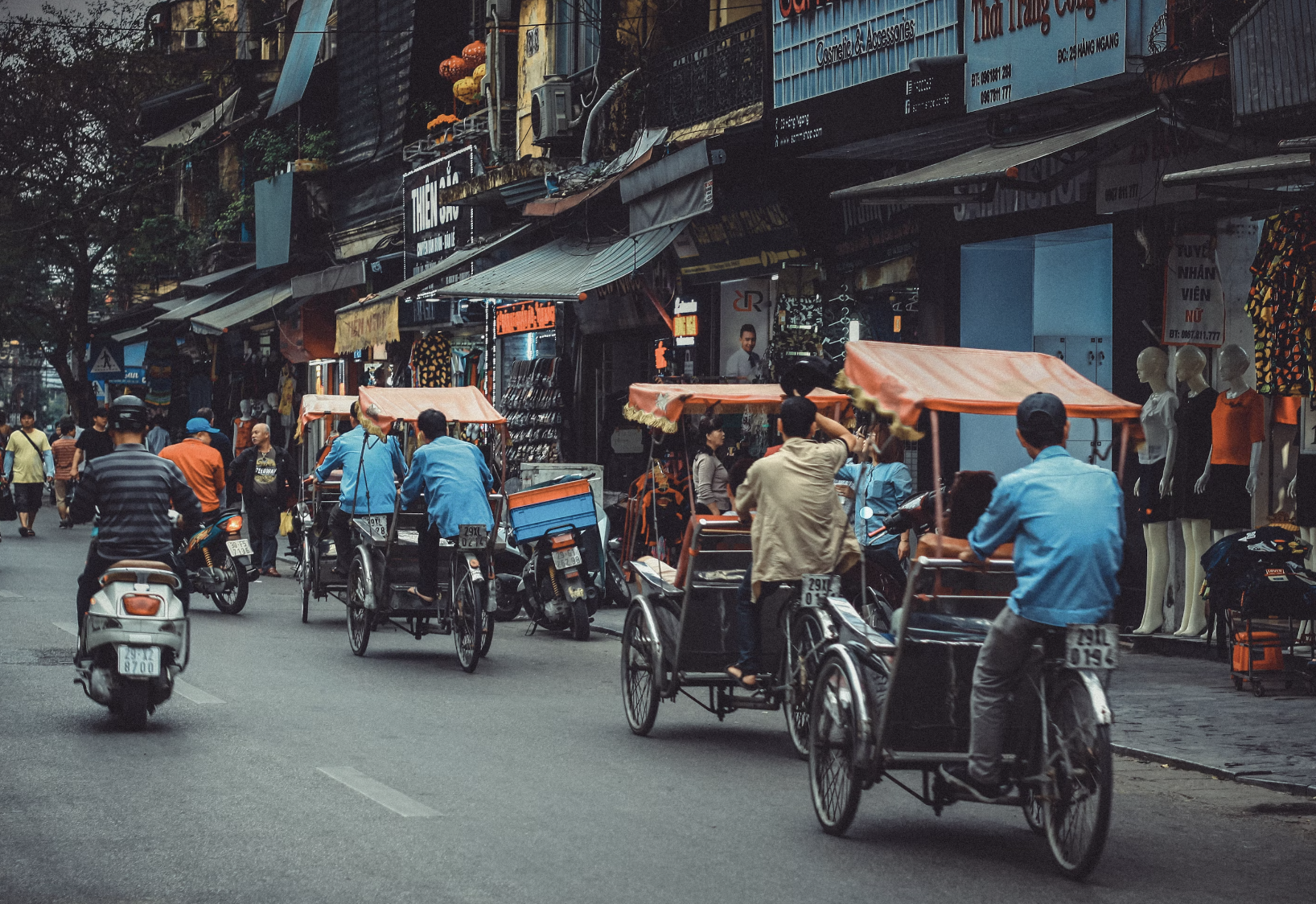 A vibrant street view of Vietnam with people riding bicycles with carriages and orange umbrellas, followed by various shops lining the road.