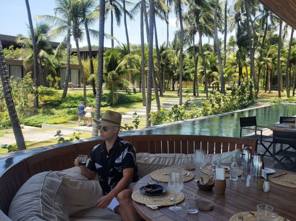 A man wearing a blue shirt and straw hat sitting on an outdoor couch next to a dining table with a swimming pool and various palm trees in the background.