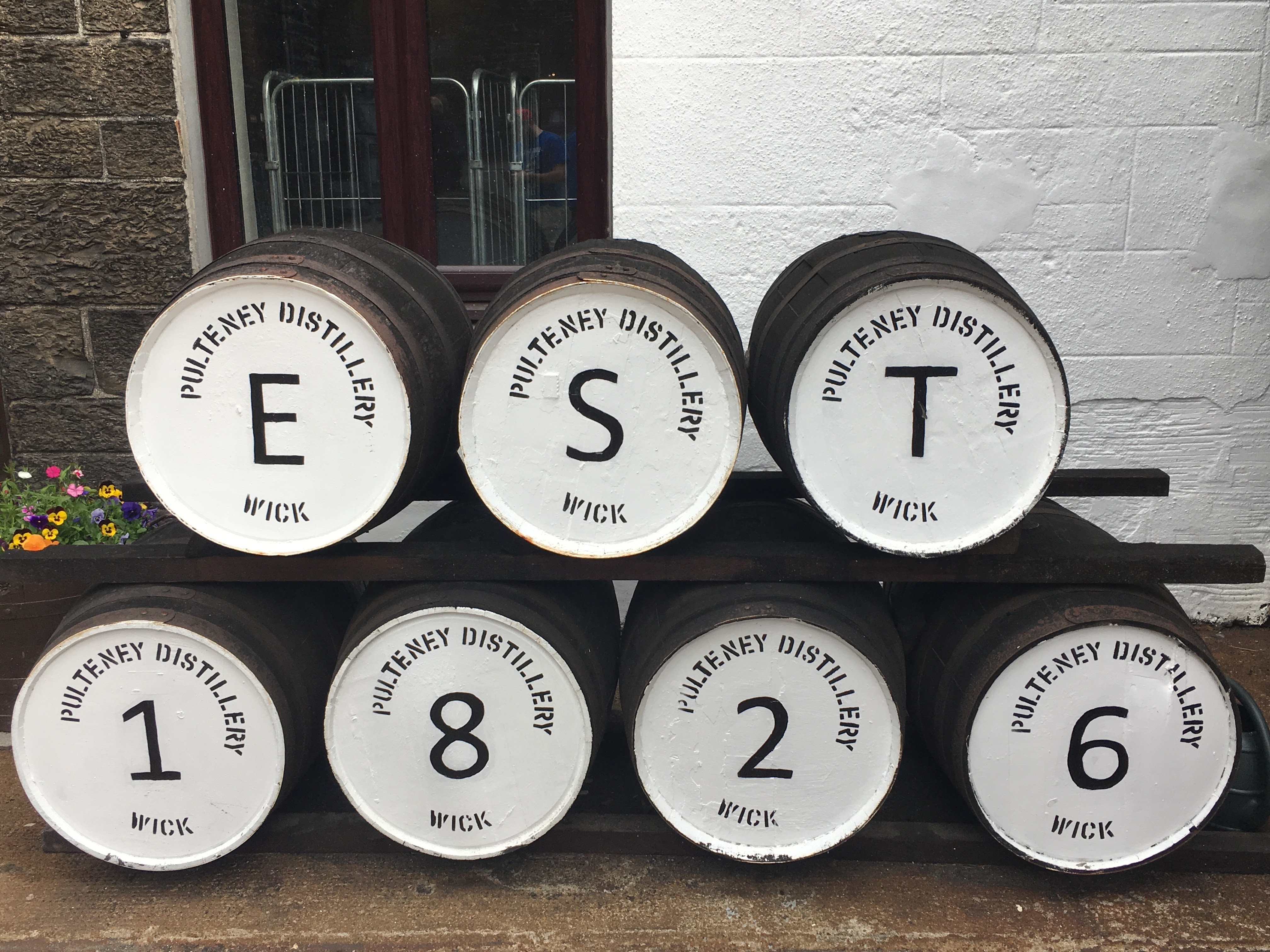 A view of seven large wine barrels painted in white that together read "EST 1826" in front of a white painted brick building and window exterior.