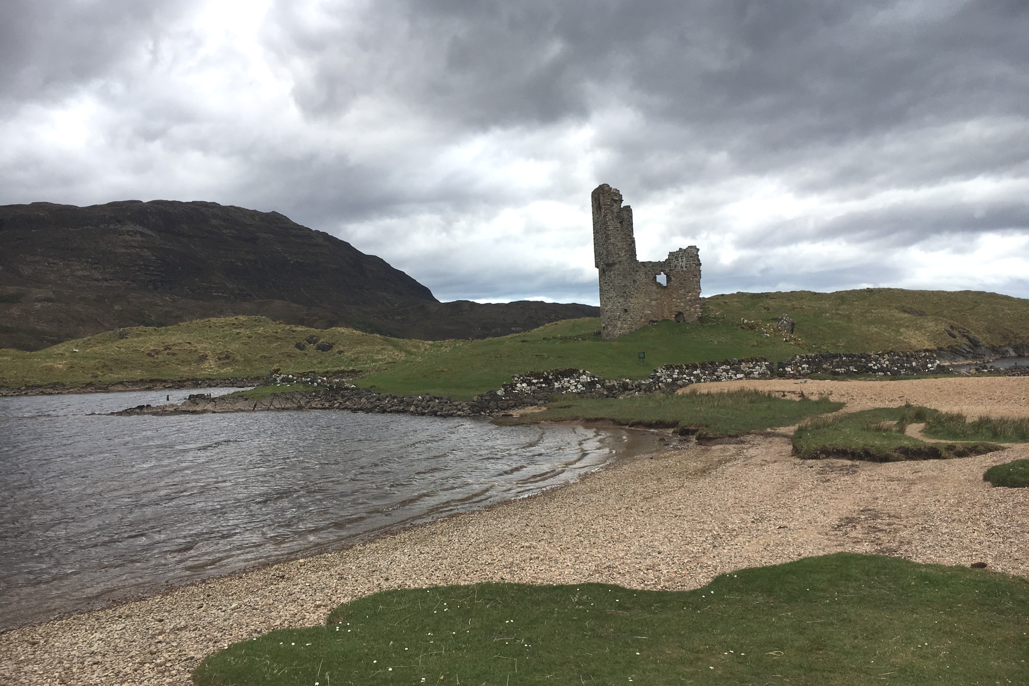 A view of stone beach surrounded by grass, water, rolling hills and a large stone castle that has deteriorated over time.