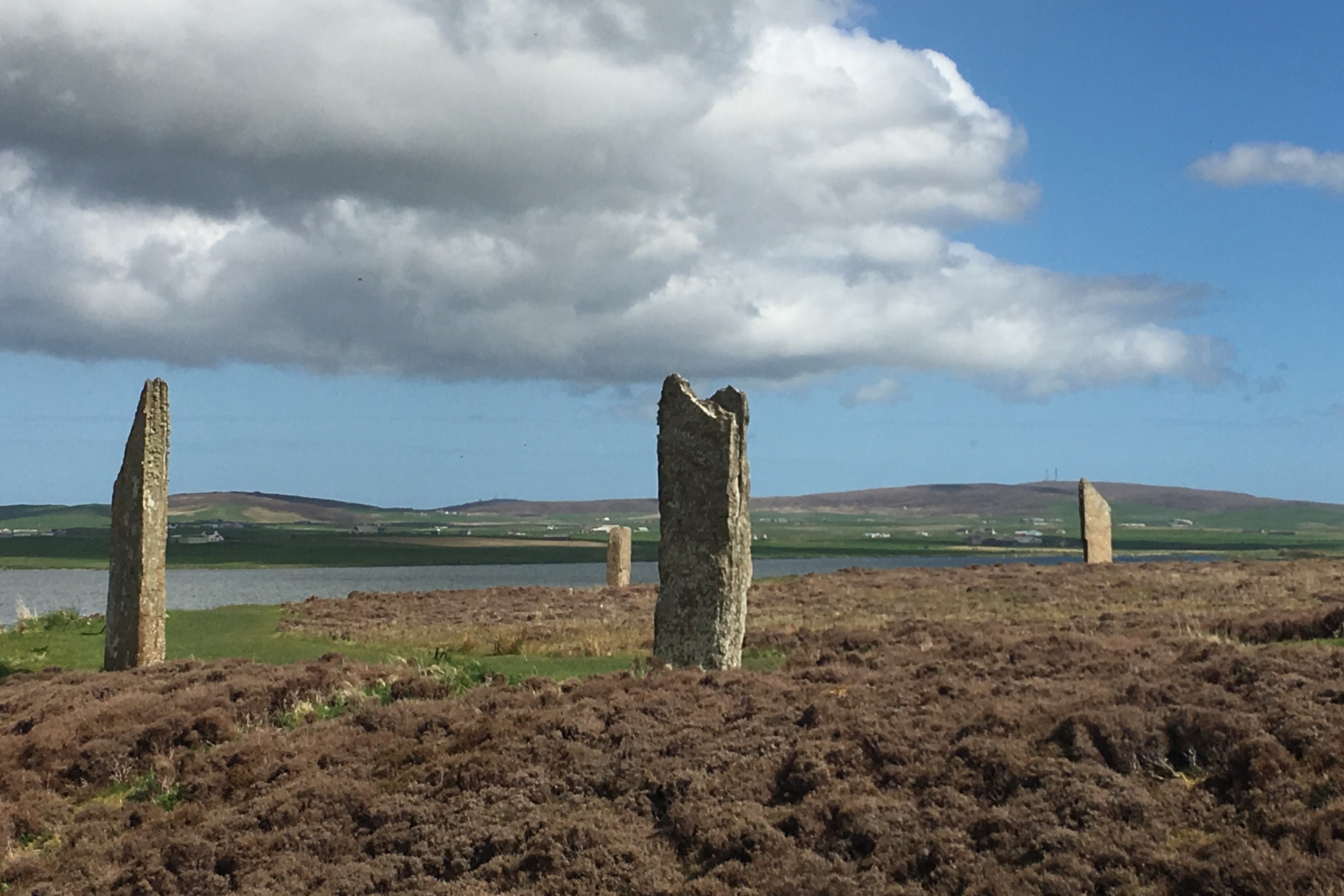 A view of four large rock pillars placed upon grassy and dirt covered terrain with rolling hills and water in the distance.