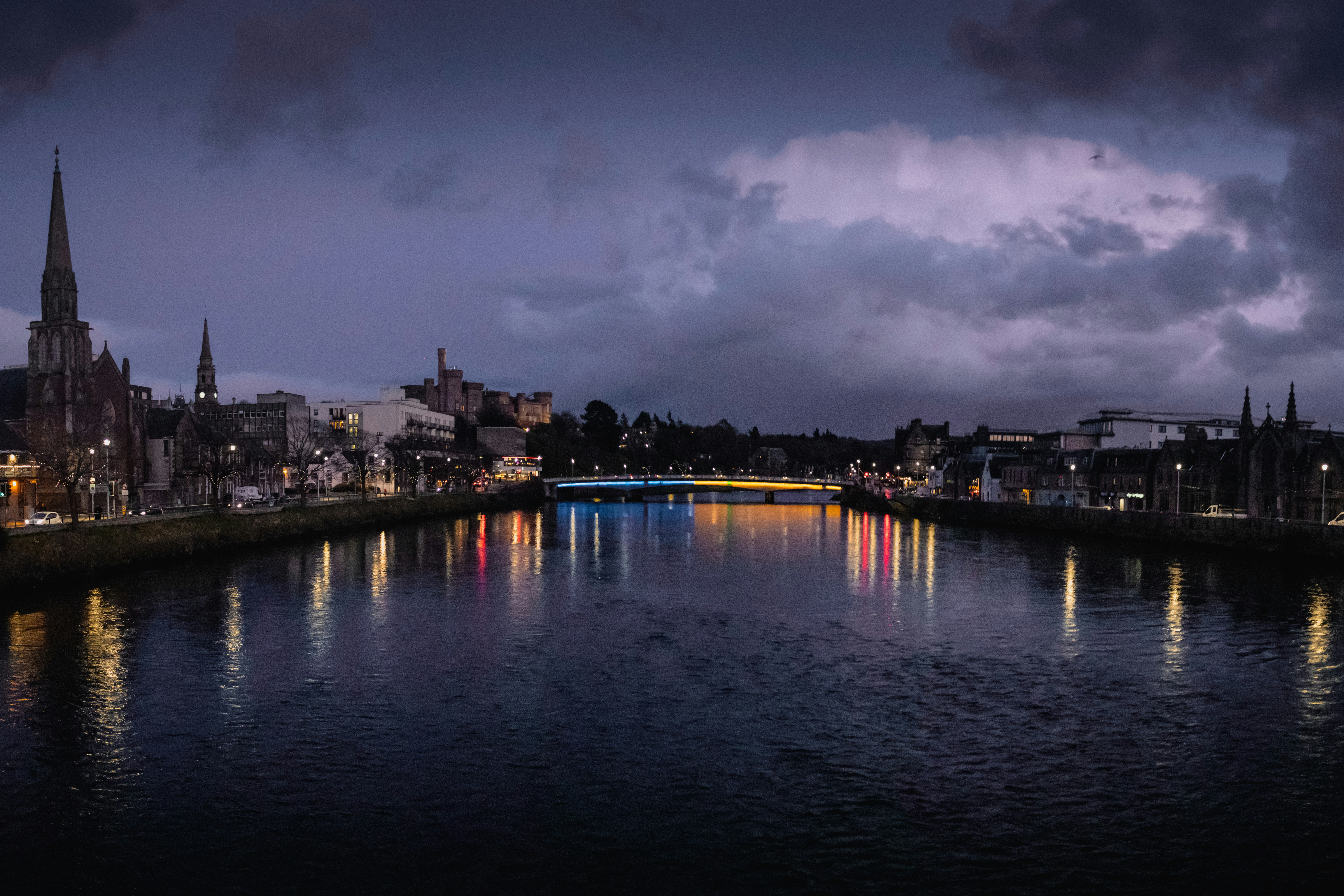 A view of a river on a cloudy night surrounded by city streets and colorful lights reflecting onto the water.