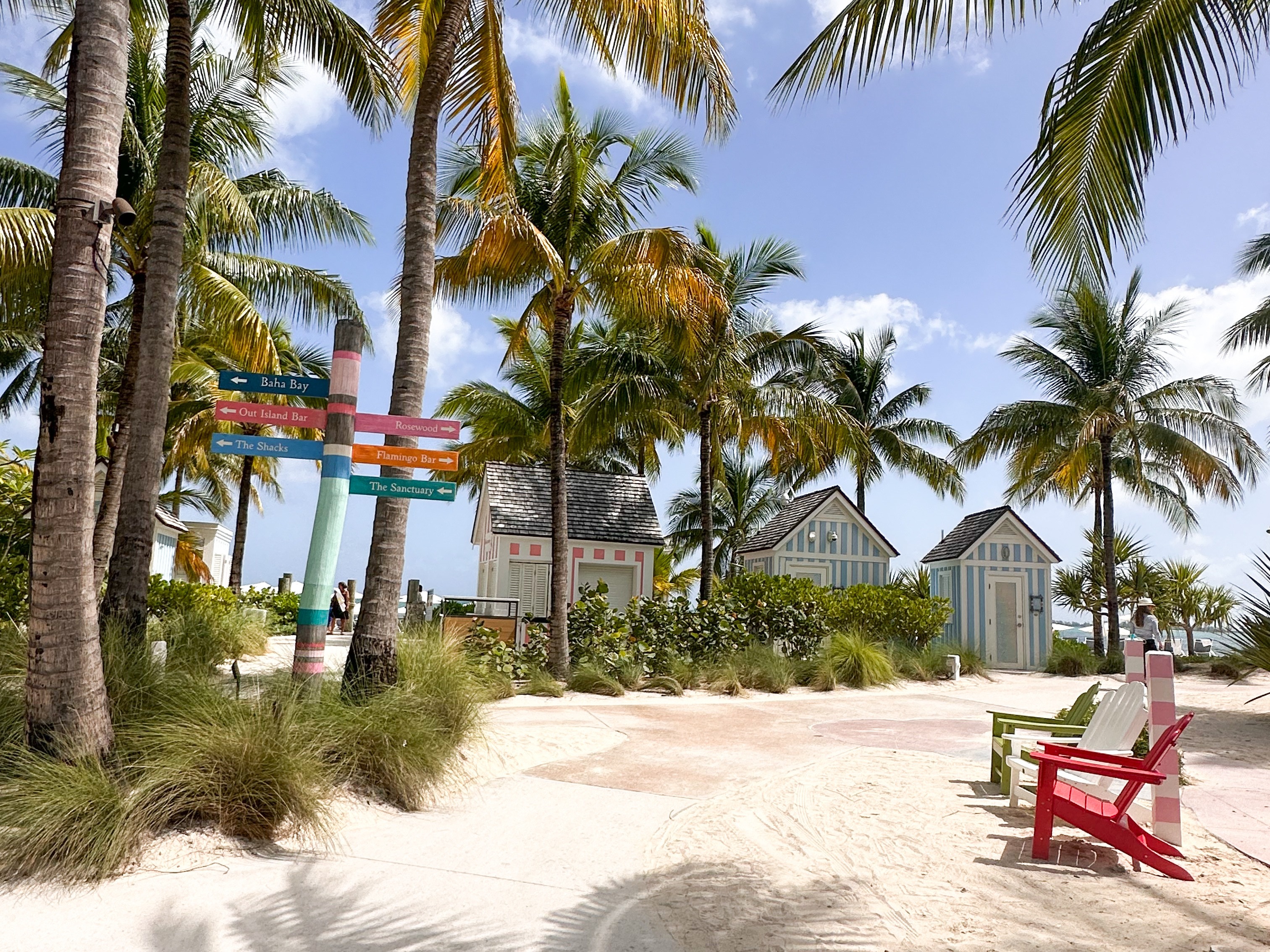 A view of a sandy white pathway surrounded by towering palm trees, a red, white and green beach chair, directional signs and three blue, white and pink striped beach shacks in the background.