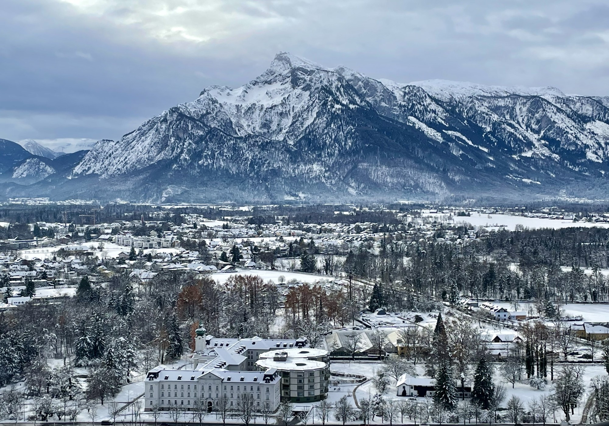 An aerial view of a hotel in a valley with houses, buildings and trees surrounding, and a large snowy mountain in the background