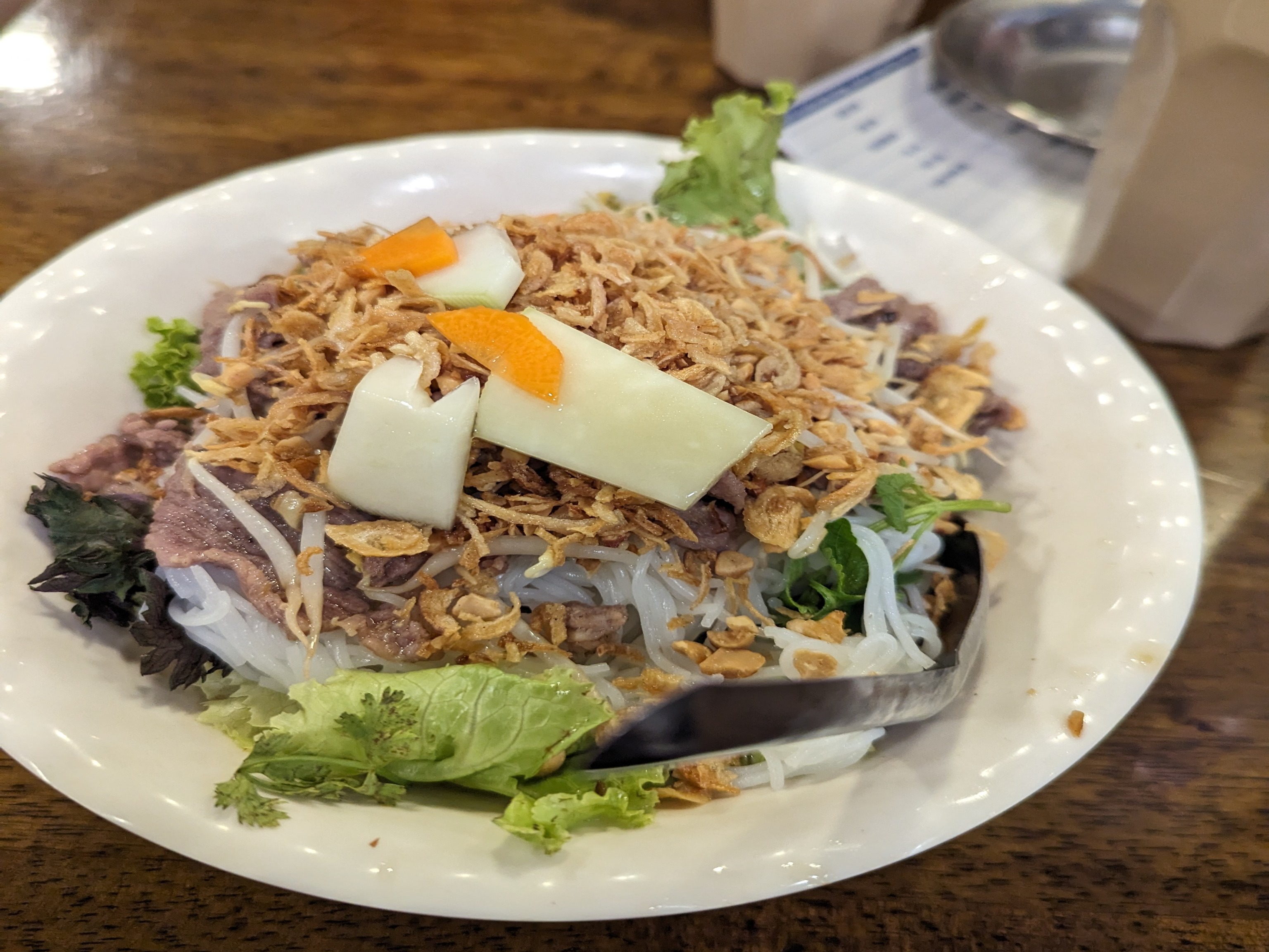 A white bowl of Vietnamese food complete with greens, noodles and various toppings placed upon a wooden table top.