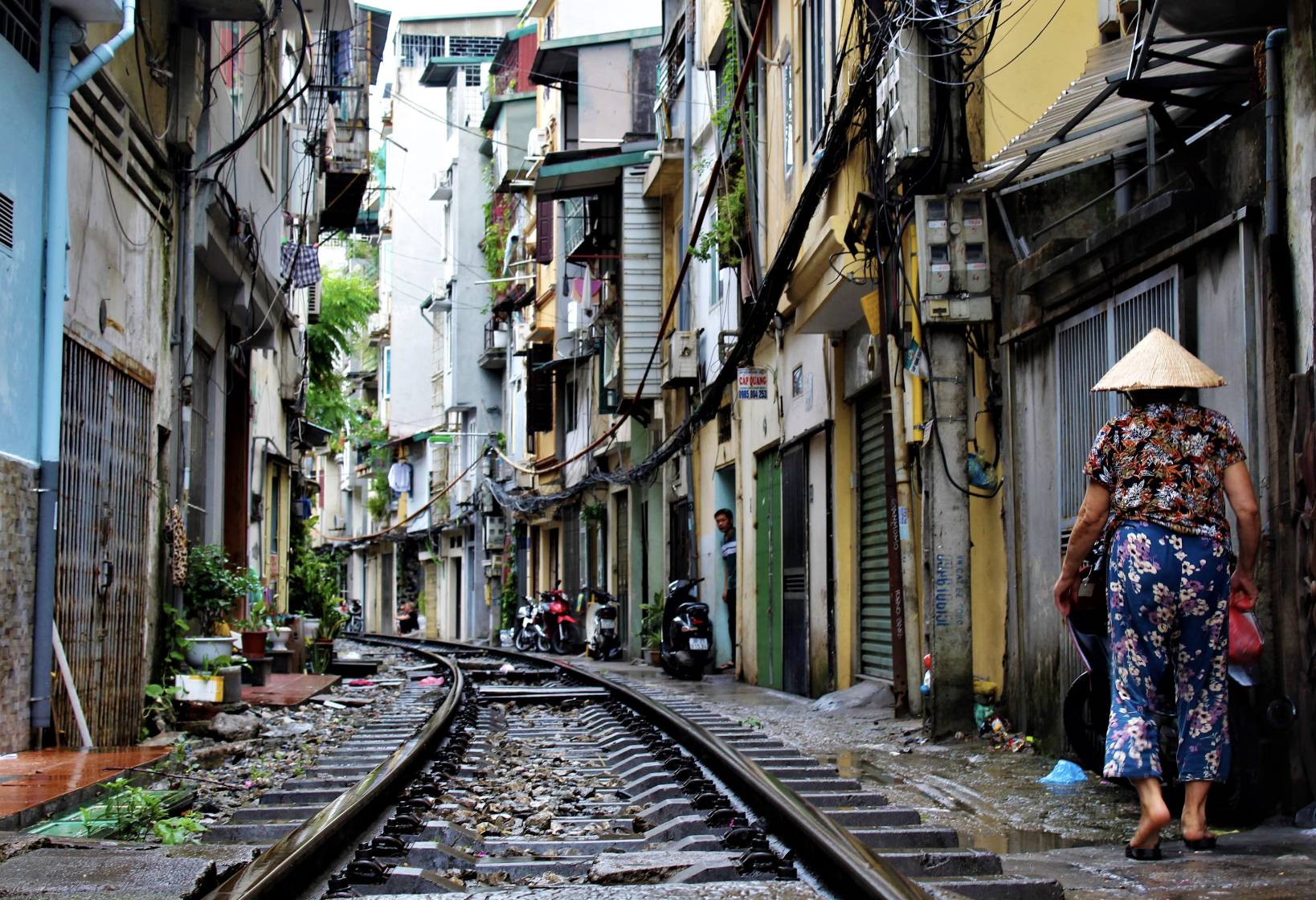 A street view of a persons back walking forward while wearing a colorfully patterned outfit and pointed straw cap, with various garages and colorful buildings in the distance alongside a railroad track and green plants scattered around.