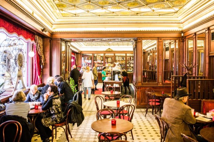 Café Maiasmokk is one of the oldest cafés in Tallinn, which has continuously been operating ever since, began in 1864. Picture is of the cafe interior, with round wooden tables, a golden ceiling, and a red-curtained window.