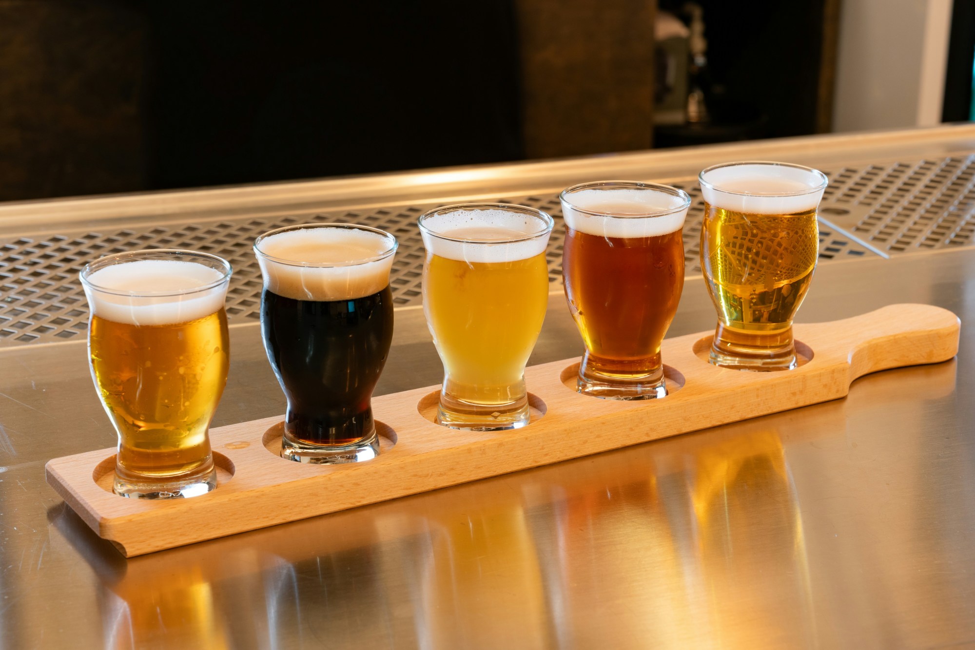 A flight of five beers, each a different shade of color lined up on a wooden board atop a stainless steel counter.