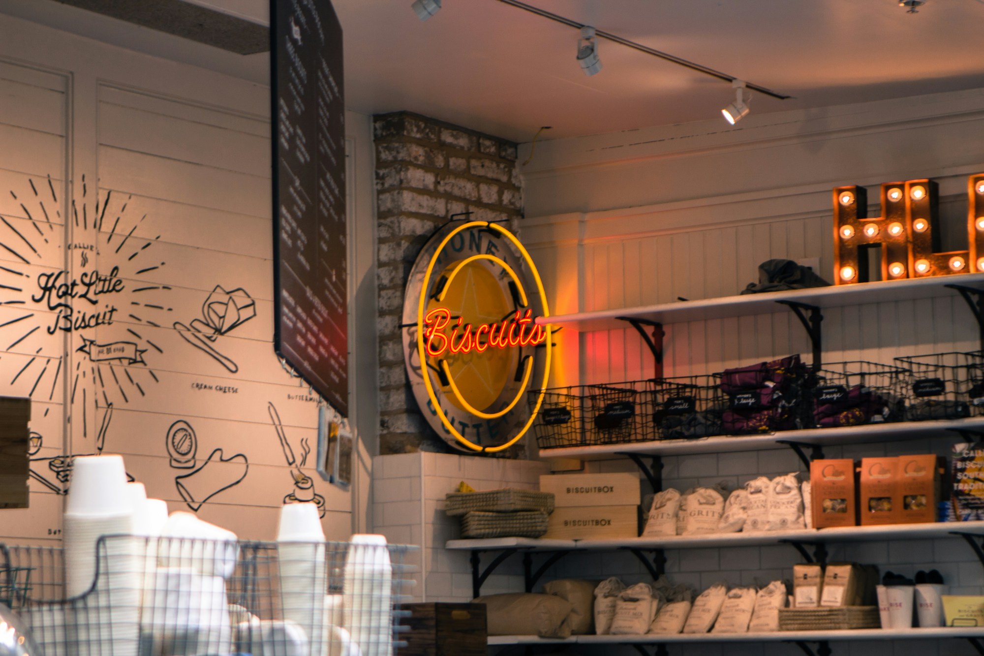 A view of a cafe with stocked supplies on a shelf, a neon sign reading 'Biscuits' in red lettering, cups in a basket and painted designs on the white paneled wall.