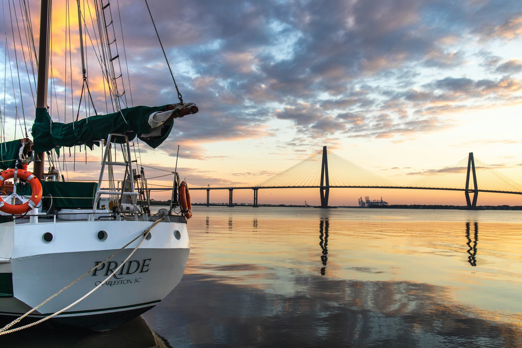 A view of a sailboat docked in a harbor in front of a bridge with a sunset and spotted clouds in the background.