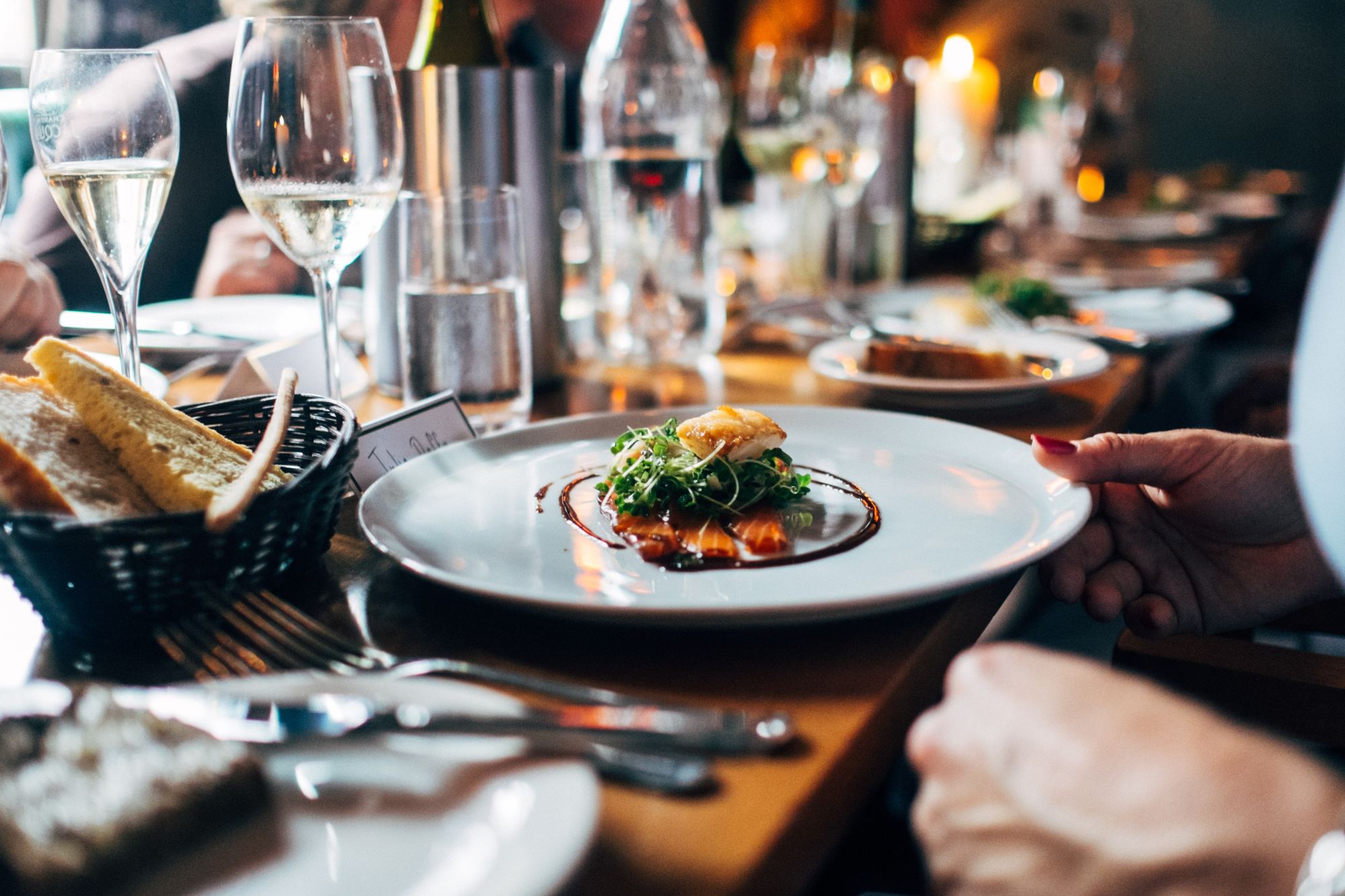 A gourmet meal plated on a white plate next to a bread basket and glass of white wine on a restaurant table with two hands nearby.