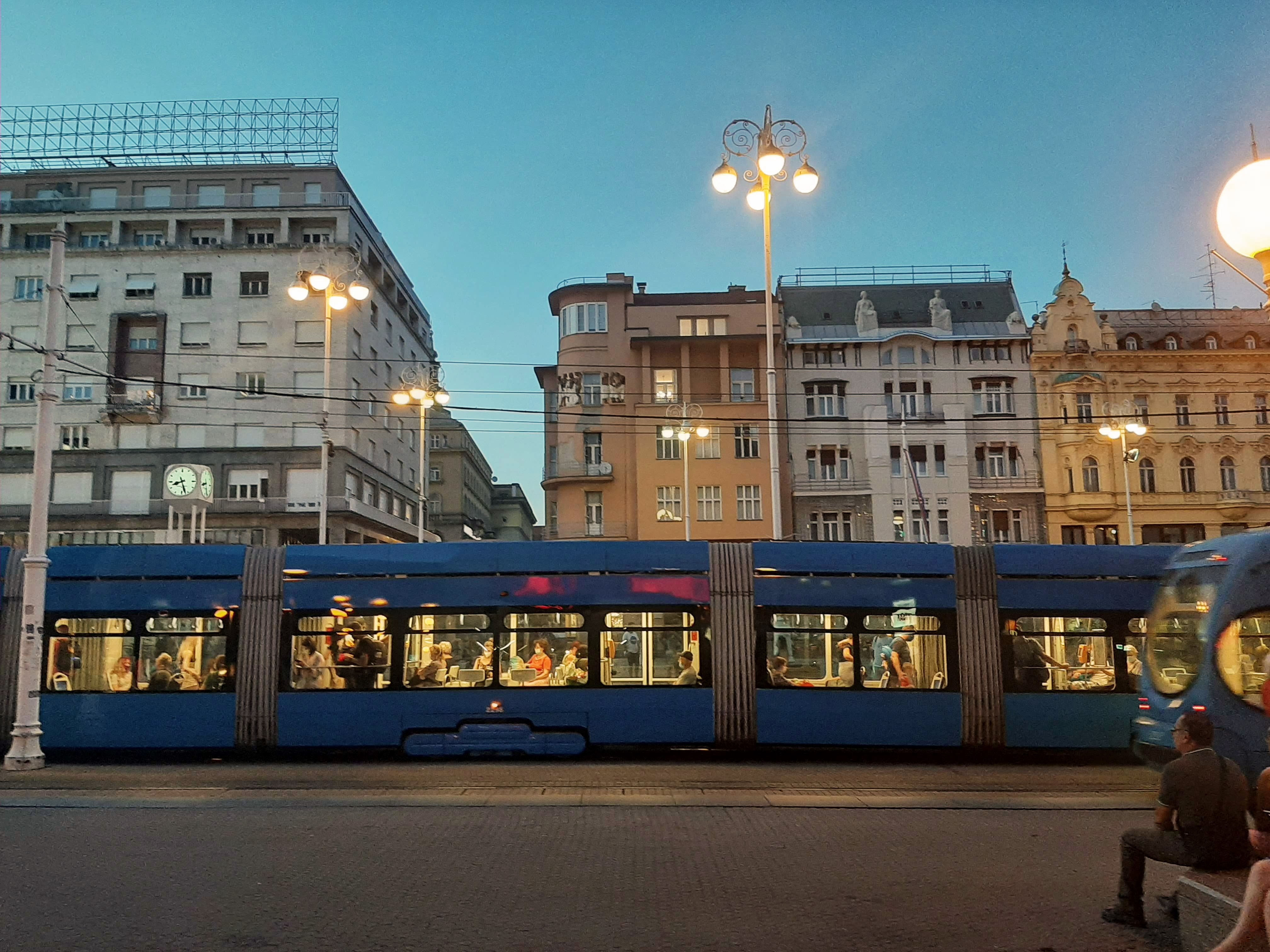 A blue train filled with passengers near the buildings during the evening time.