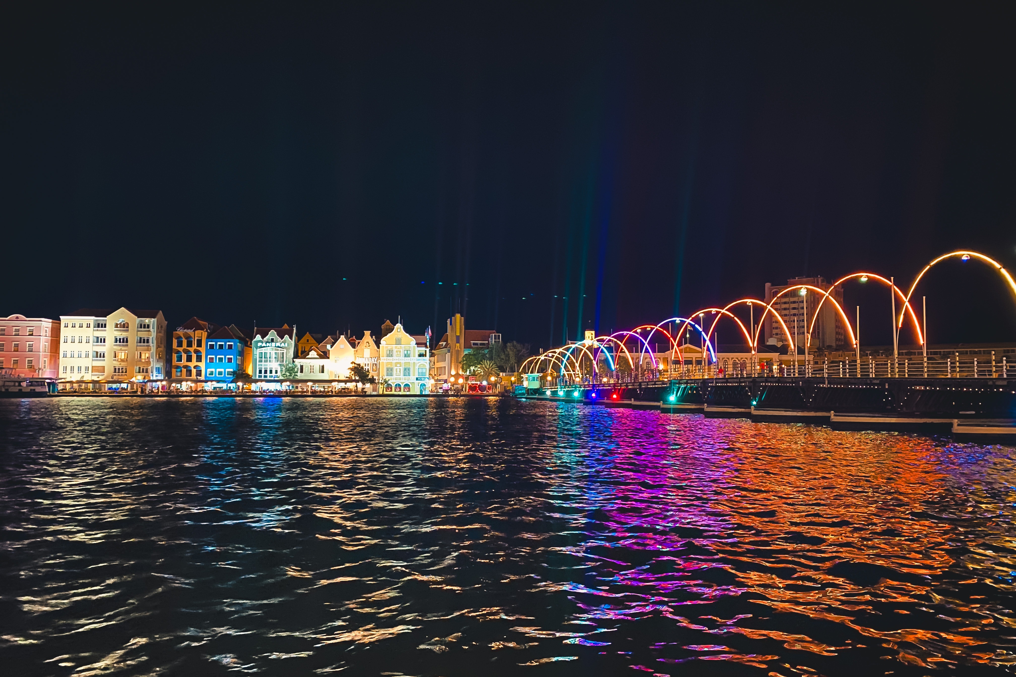 The Queen Emma Bridge, also known as the "Swinging Old Lady," during nighttime lit up with multiple colors reflecting against the water.