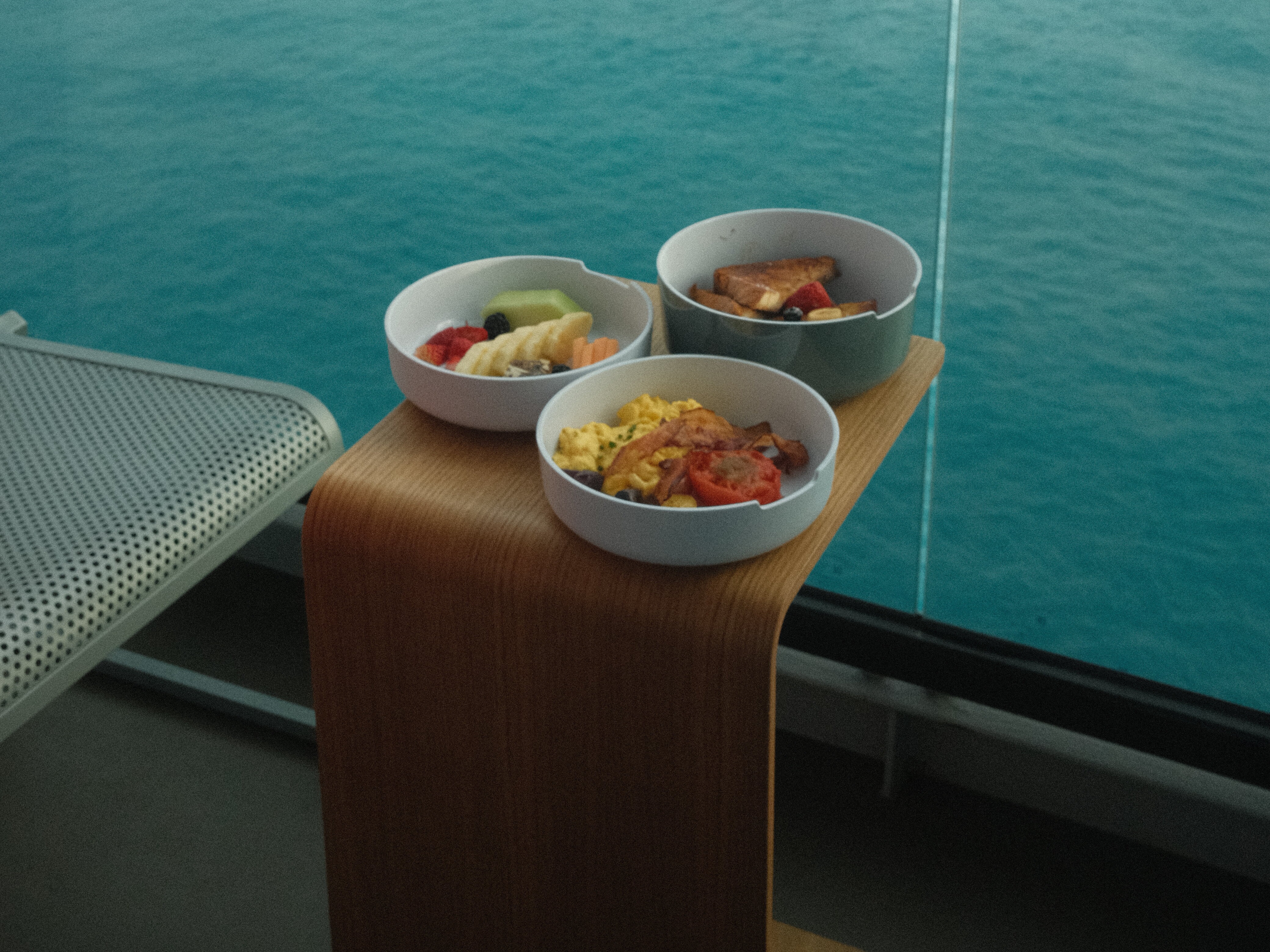 A picture of bowls of fruits on a wooden table near the sea.