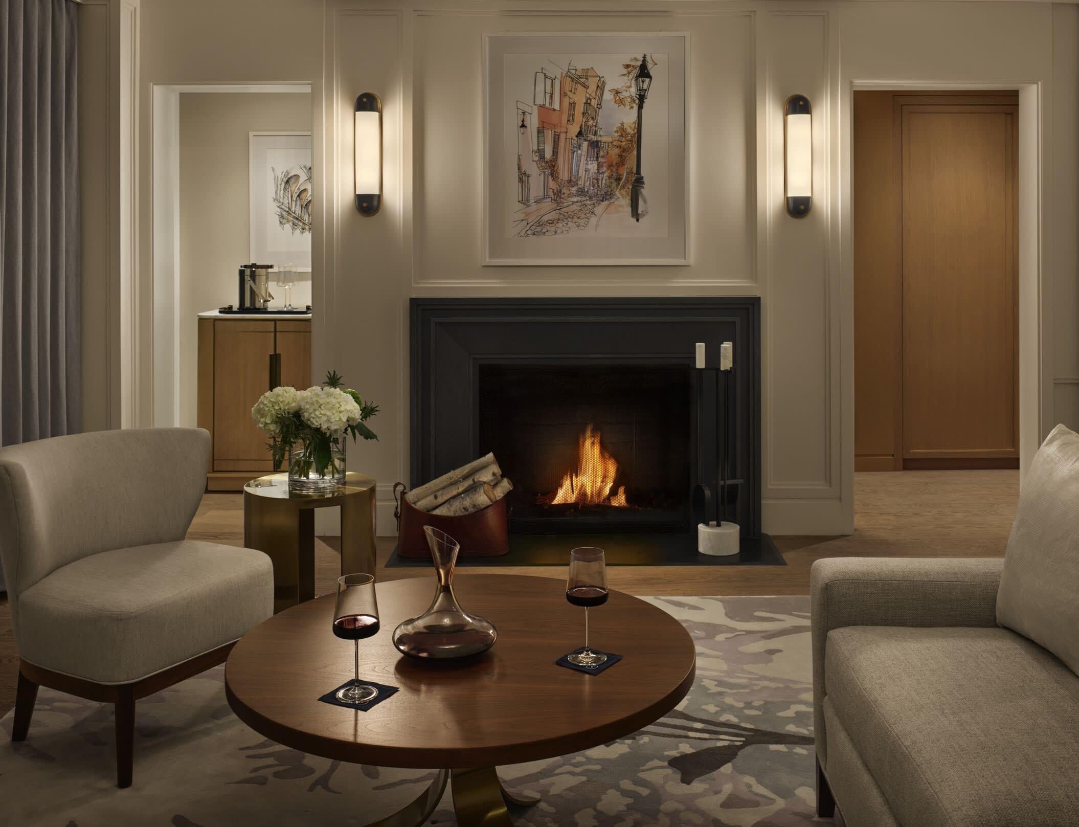 Elegant hotel suite with a cozy fireplace, modern armchairs, fresh flowers, and wine glasses on a wooden coffee table.