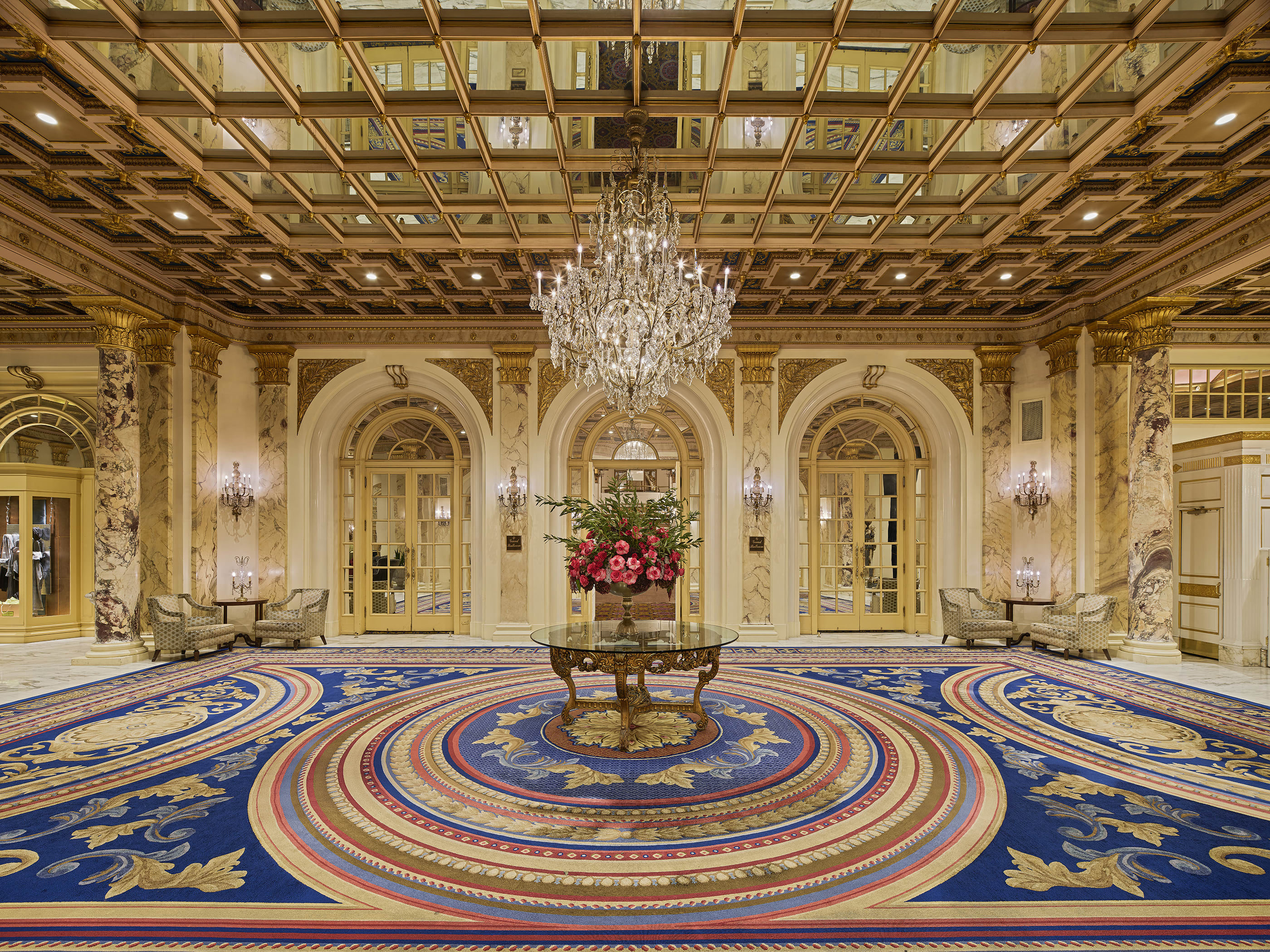 Grand hotel lobby with ornate marble walls, gilded accents, and a mirrored coffered ceiling featuring a large crystal chandelier. A round table with a floral arrangement sits at the center of an elaborate blue, gold, and red patterned carpet.