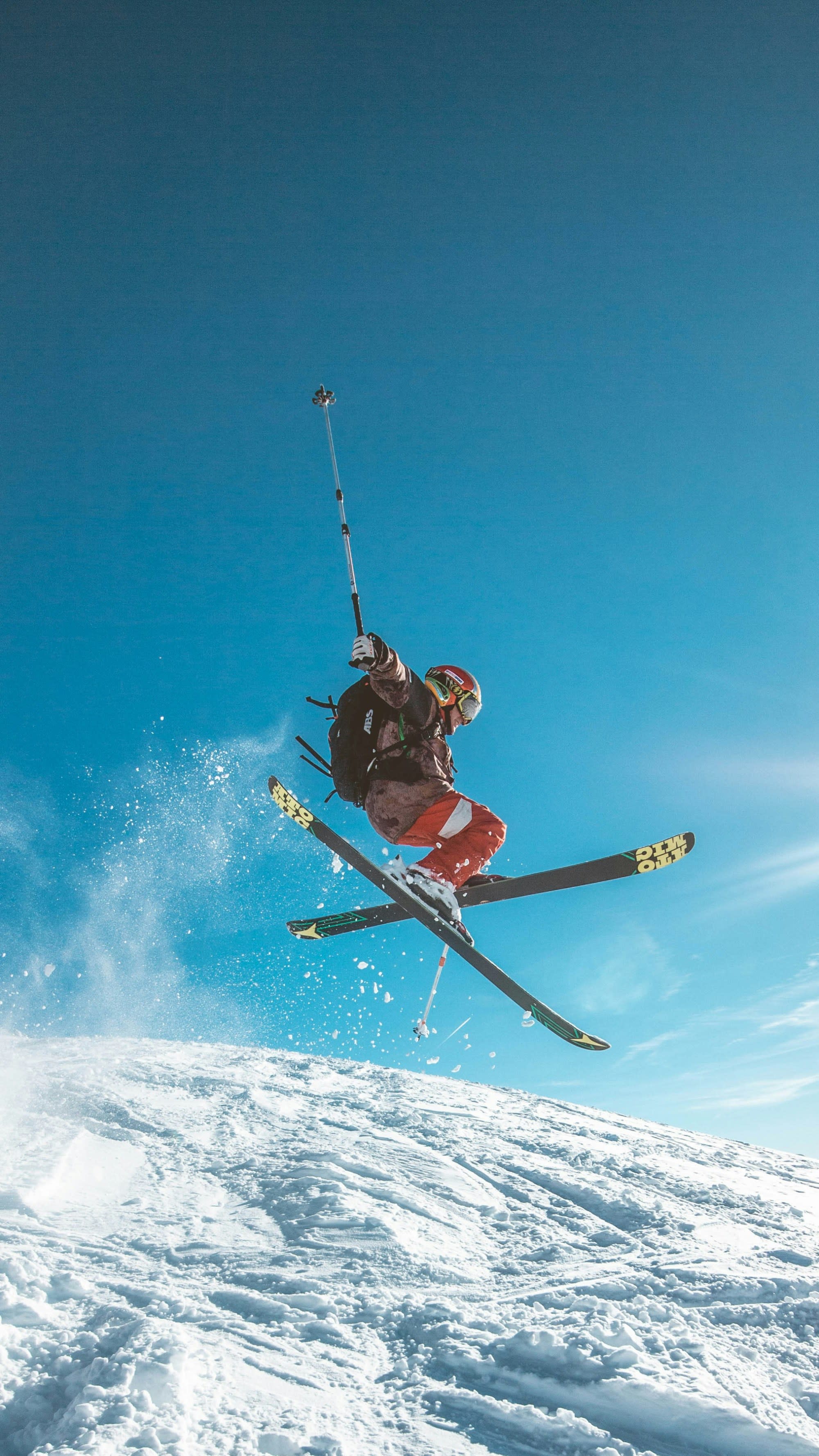 A man on skis jumps above the crest of a snowy hill with a clear blue sky behind.