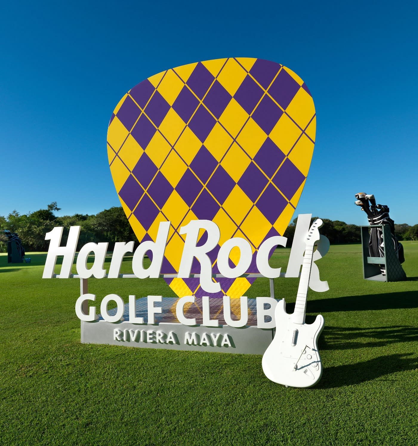 A large sign on a gold course reading "Hard Rock Golf Club Riviera Maya" with a yellow and purple guitar pick statue behind it and a white guitar in front