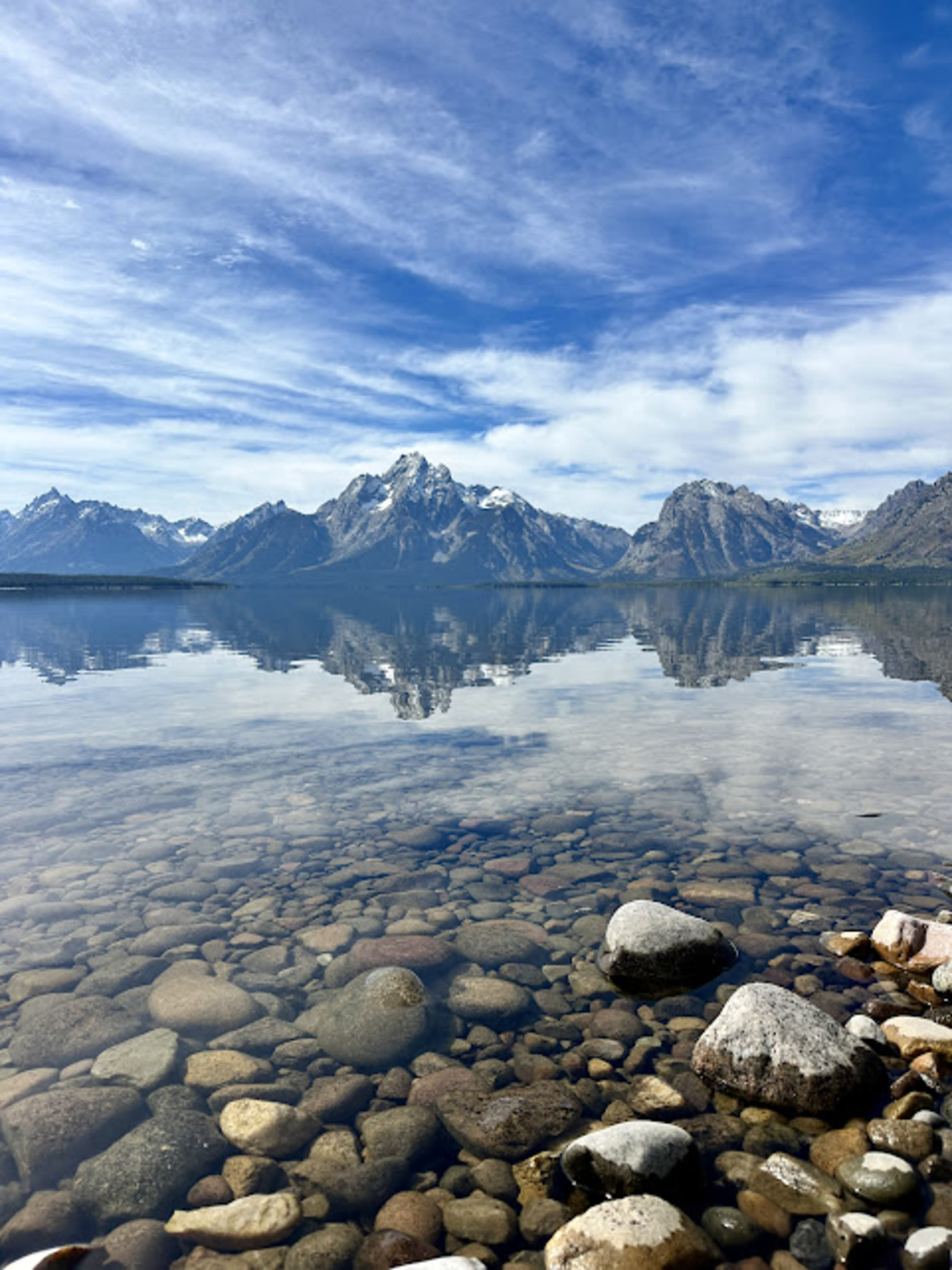 A clear lake reflects a mountain range under a blue sky with scattered clouds, with visible stones beneath the water in the foreground.