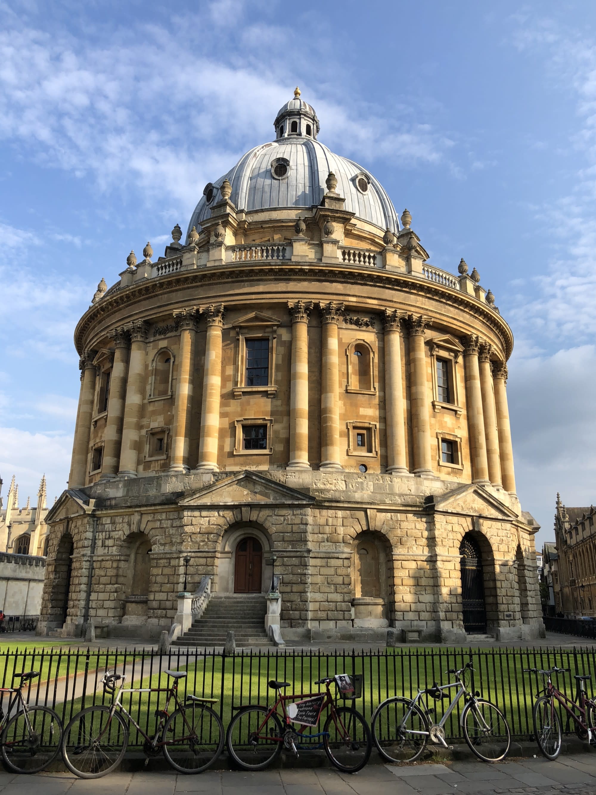 A view of a large dome building with ornate architexture and an iron gate surrounding it, known as the Radcliffe Camera, during the day.