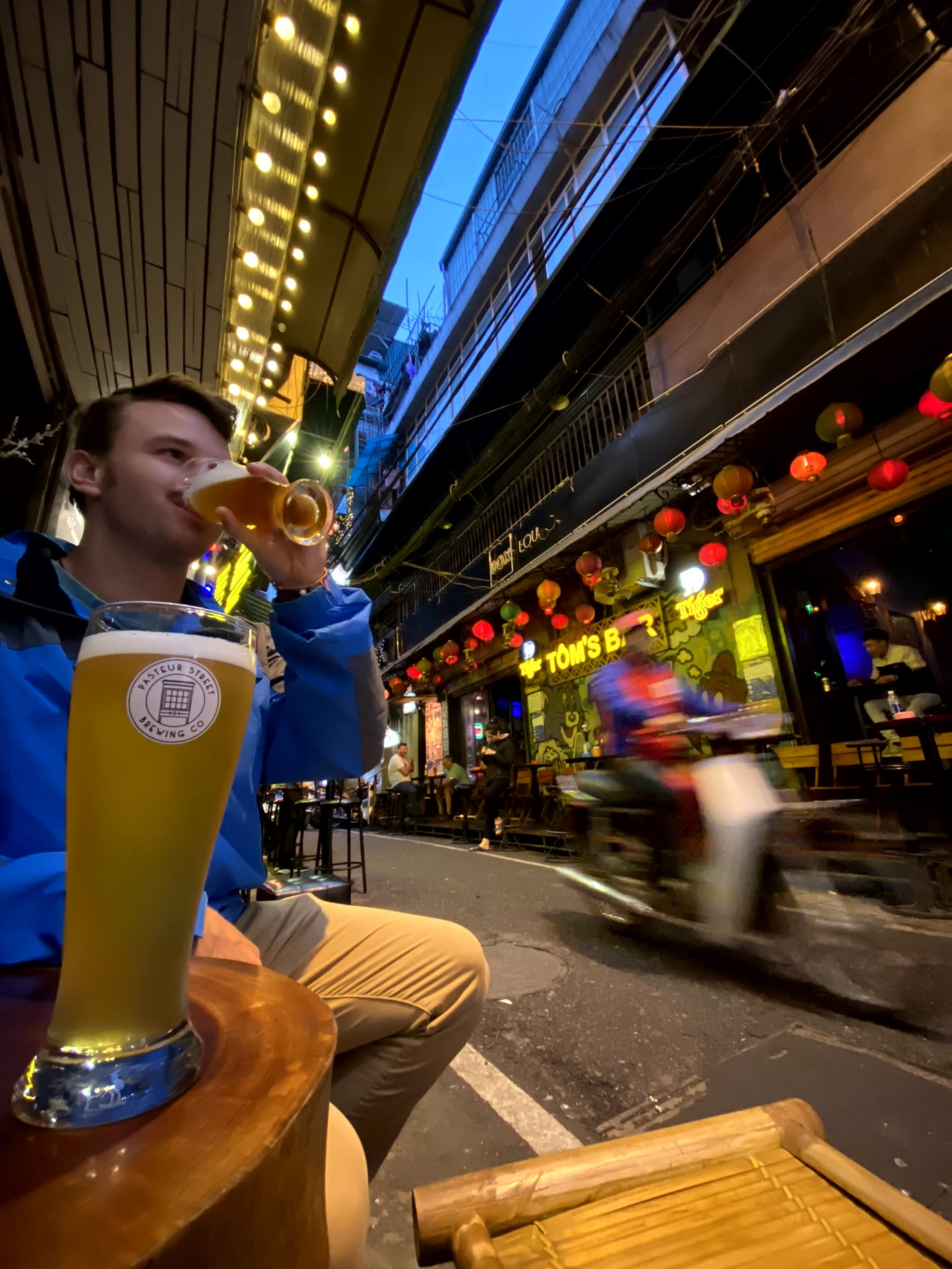 A blurred urban street scene at night, viewed from above, with a focus on a person’s hand holding a beverage.