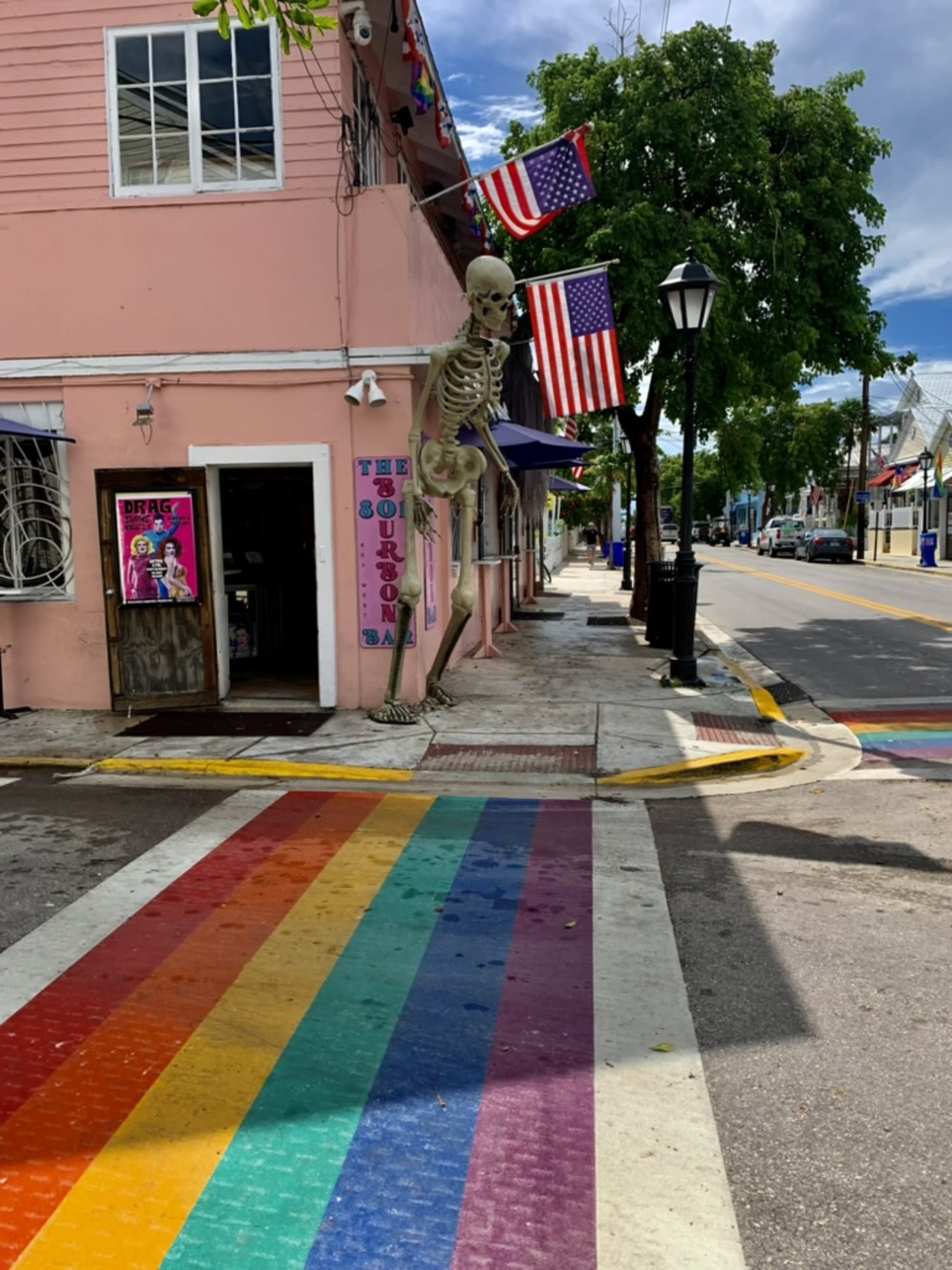 The image shows a vibrant street corner with a rainbow-colored crosswalk, a pink building adorned with American flags, a skeleton decoration, and a lamppost.