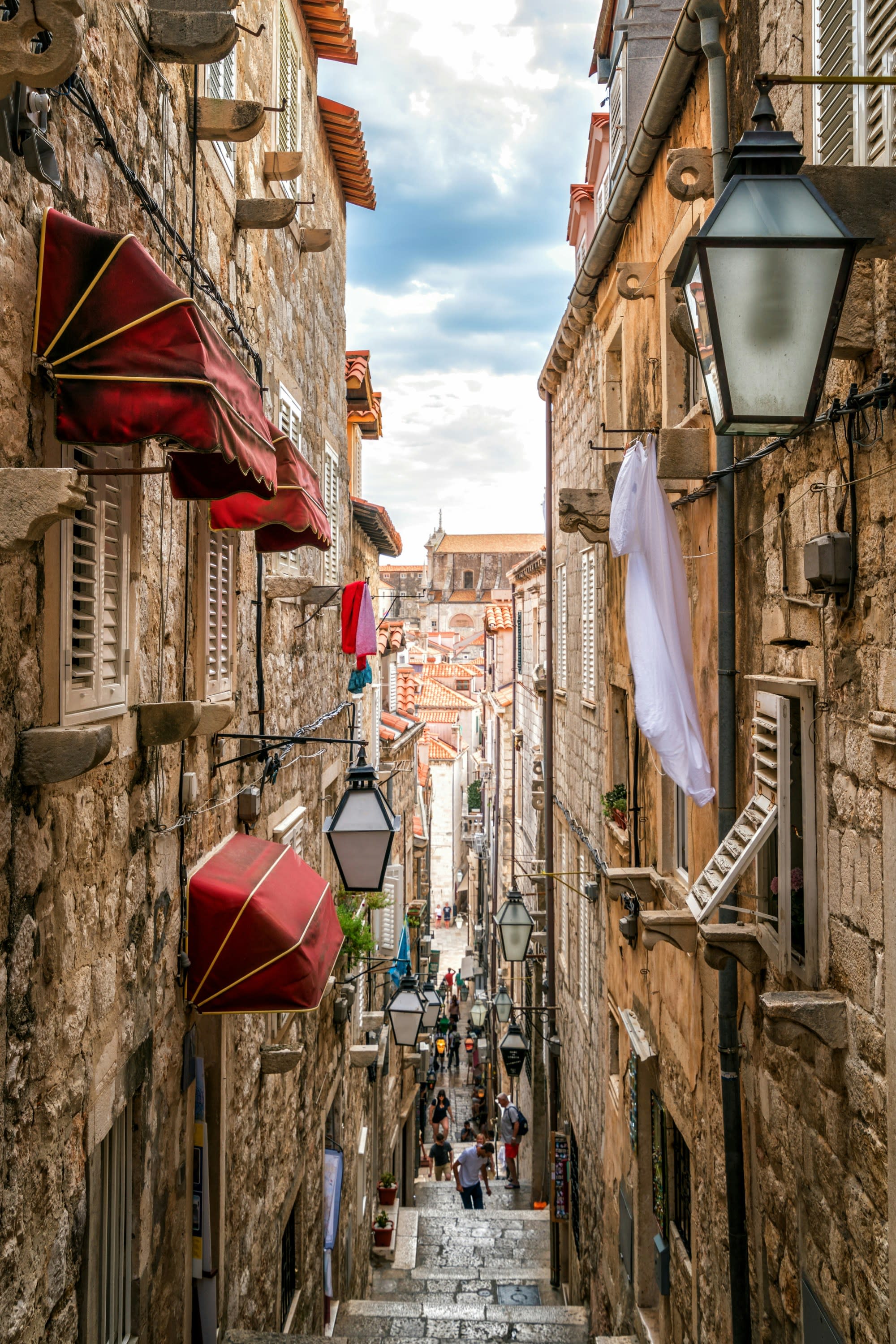 A narrow, stone-paved street in a historic European city, bustling with people and lined with old buildings and hanging lanterns.