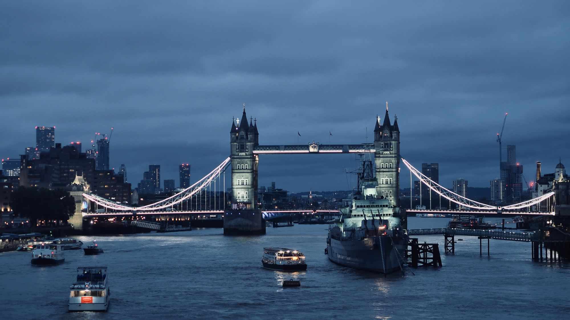 Night time cloudy city skyline with bridge , water body with boats and buildings in the background.