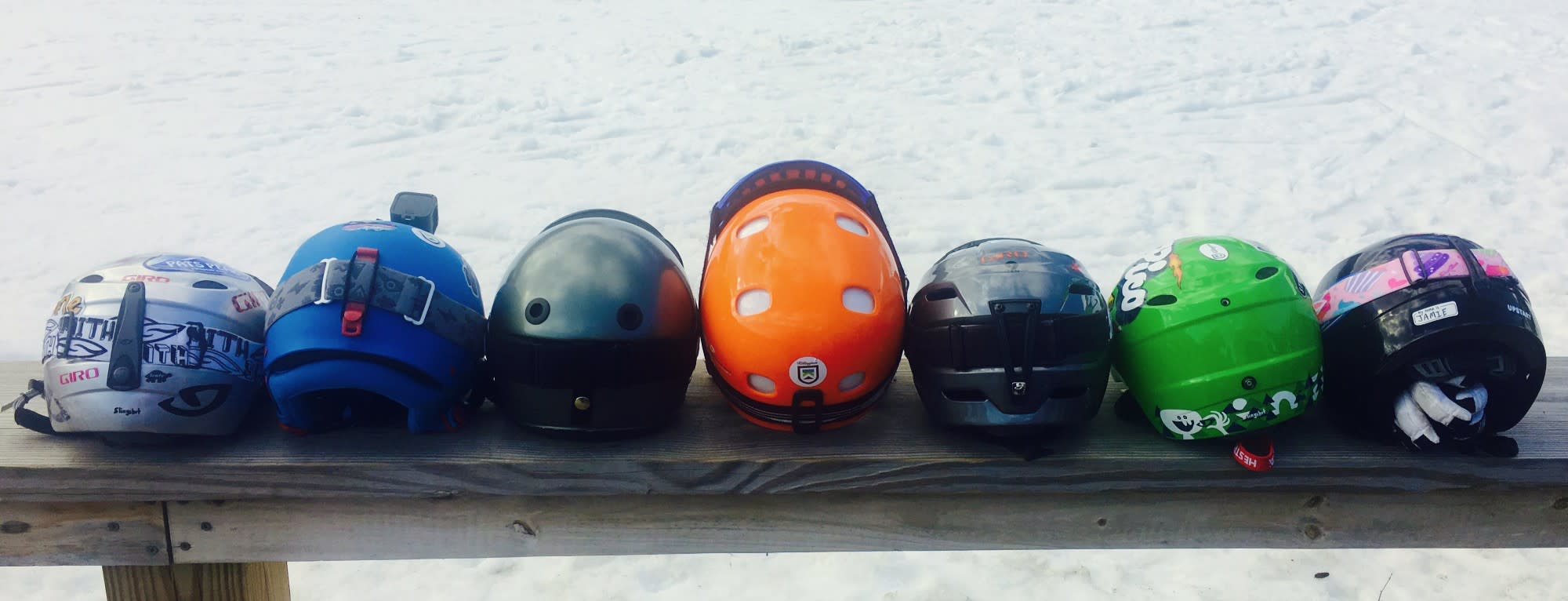 A picture of seven different colored ski helmets lined up on a wooden bench.