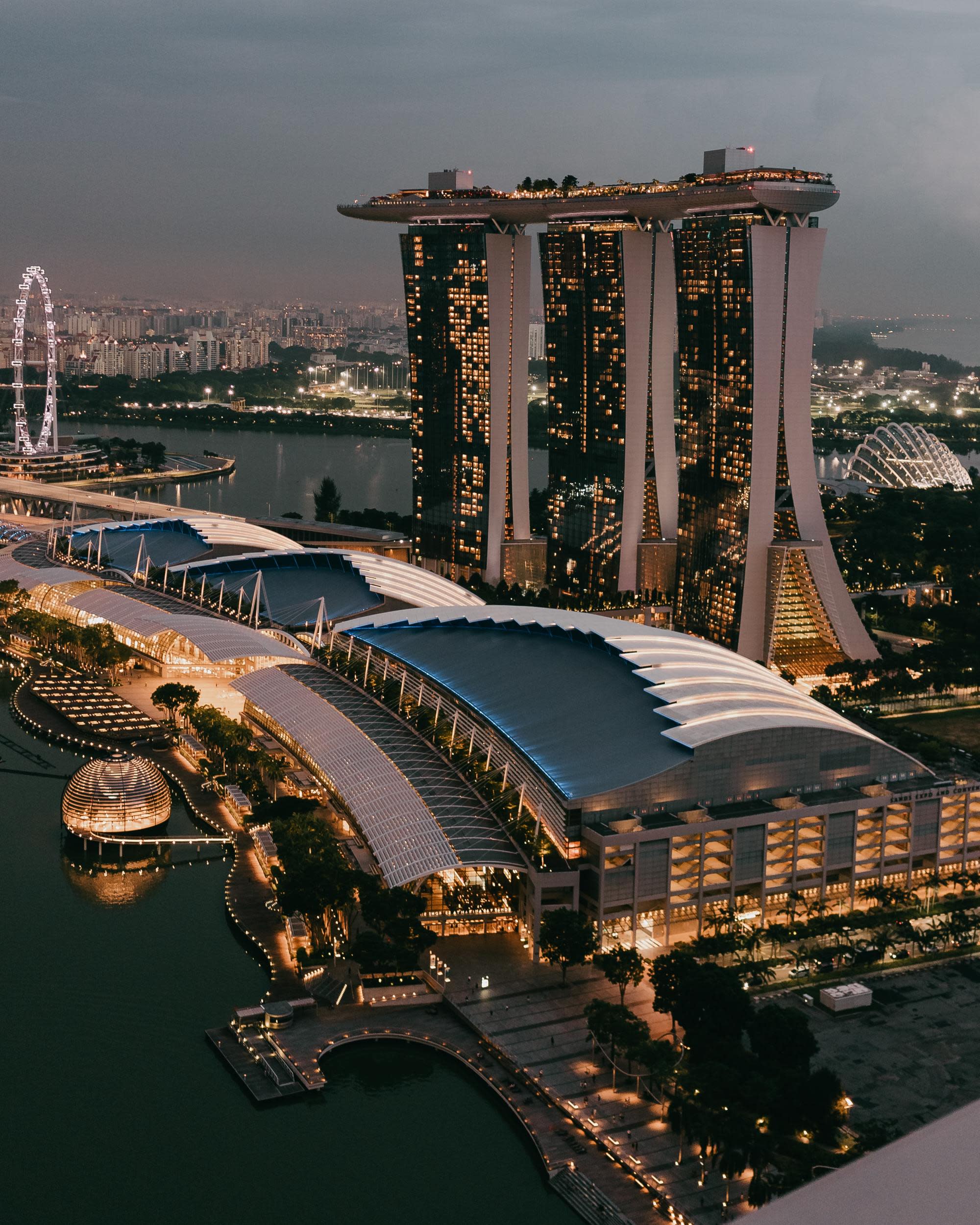 three tall buildings lit up at night on the water surrounded by sprawling modern city