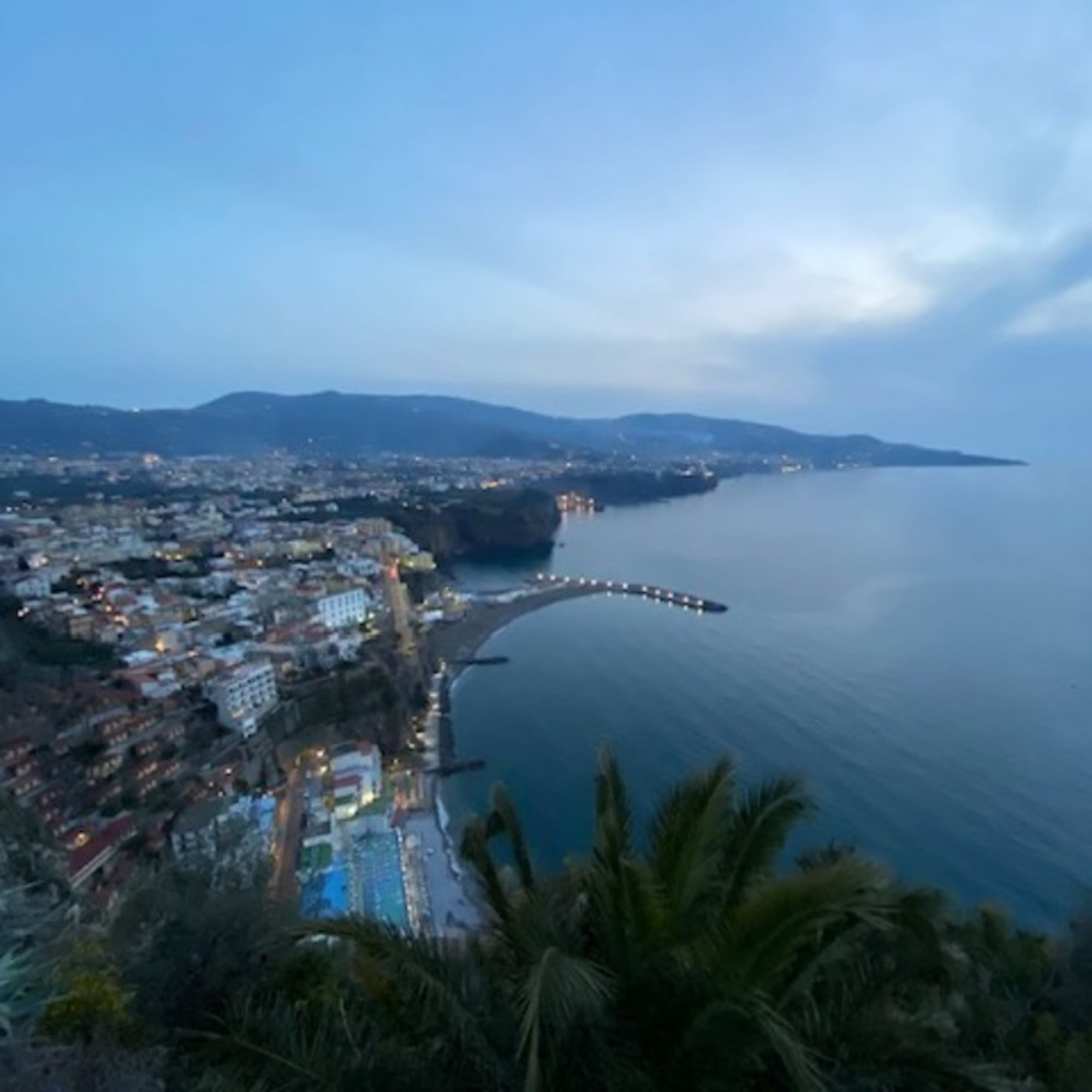 The image depicts a coastal cityscape during twilight with lights beginning to illuminate the buildings and a curved pier extending into the calm sea, all viewed from an elevated vantage point.