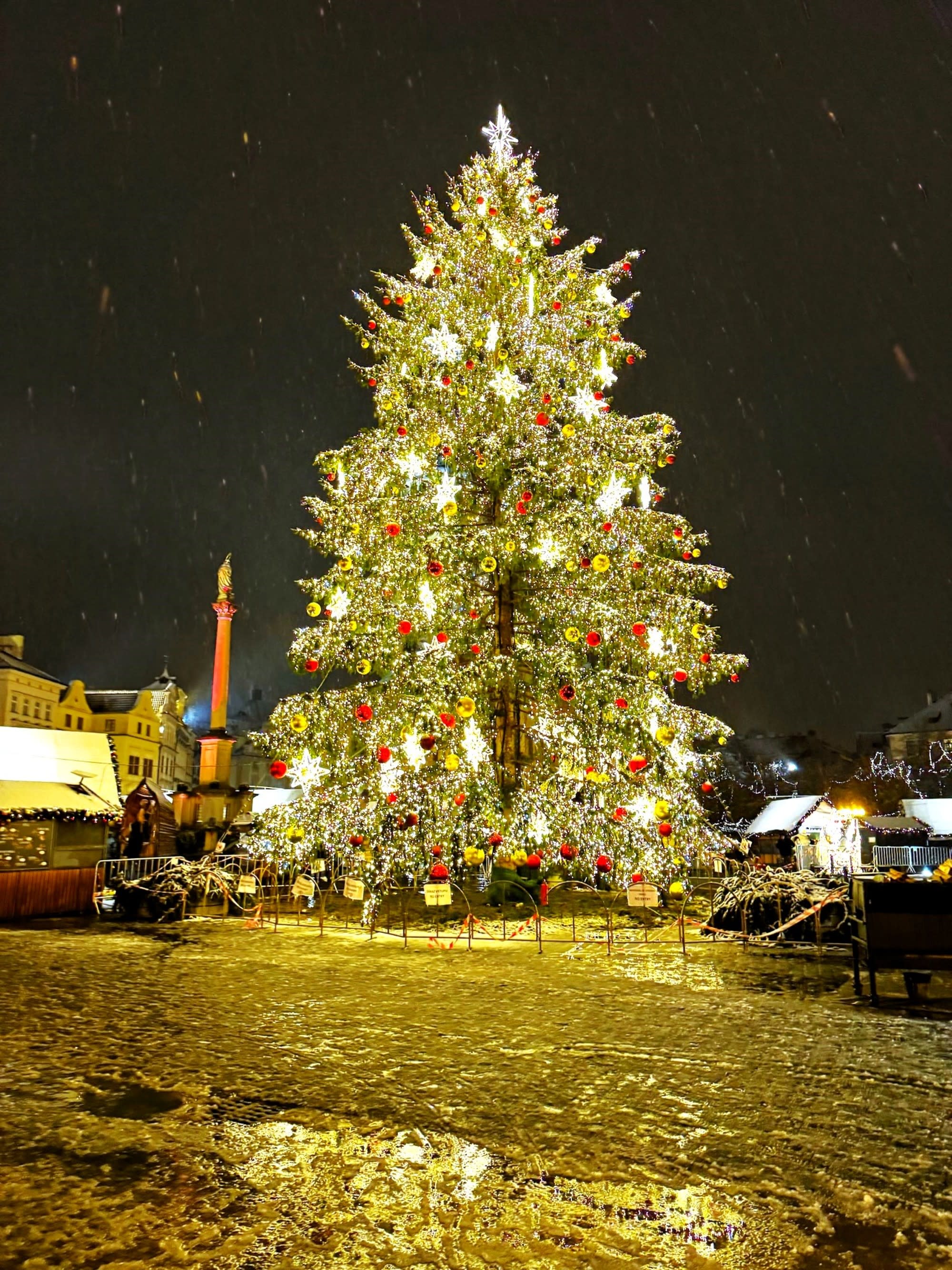 A large illuminated Christmas tree adorned with red ornaments and lights, set in a nighttime outdoor setting with snowfall in Prague.