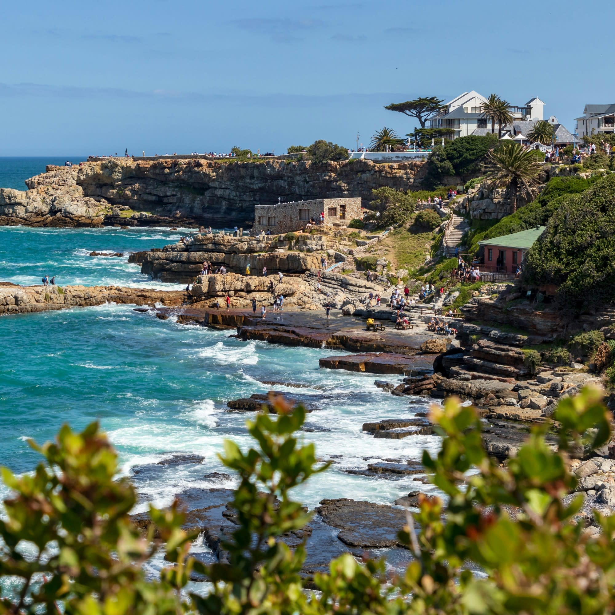 The image depicts a rocky coastline with waves crashing against the shore, vegetation in the foreground, and buildings on the cliff edge under a clear sky.