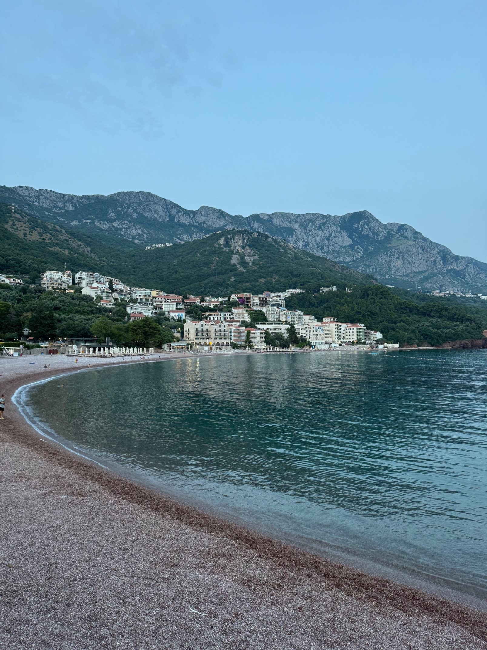 A scenic view of calm waves washing onto a beach with a coastal town in the disance and mountains at Kotor Bay, Montenegro.