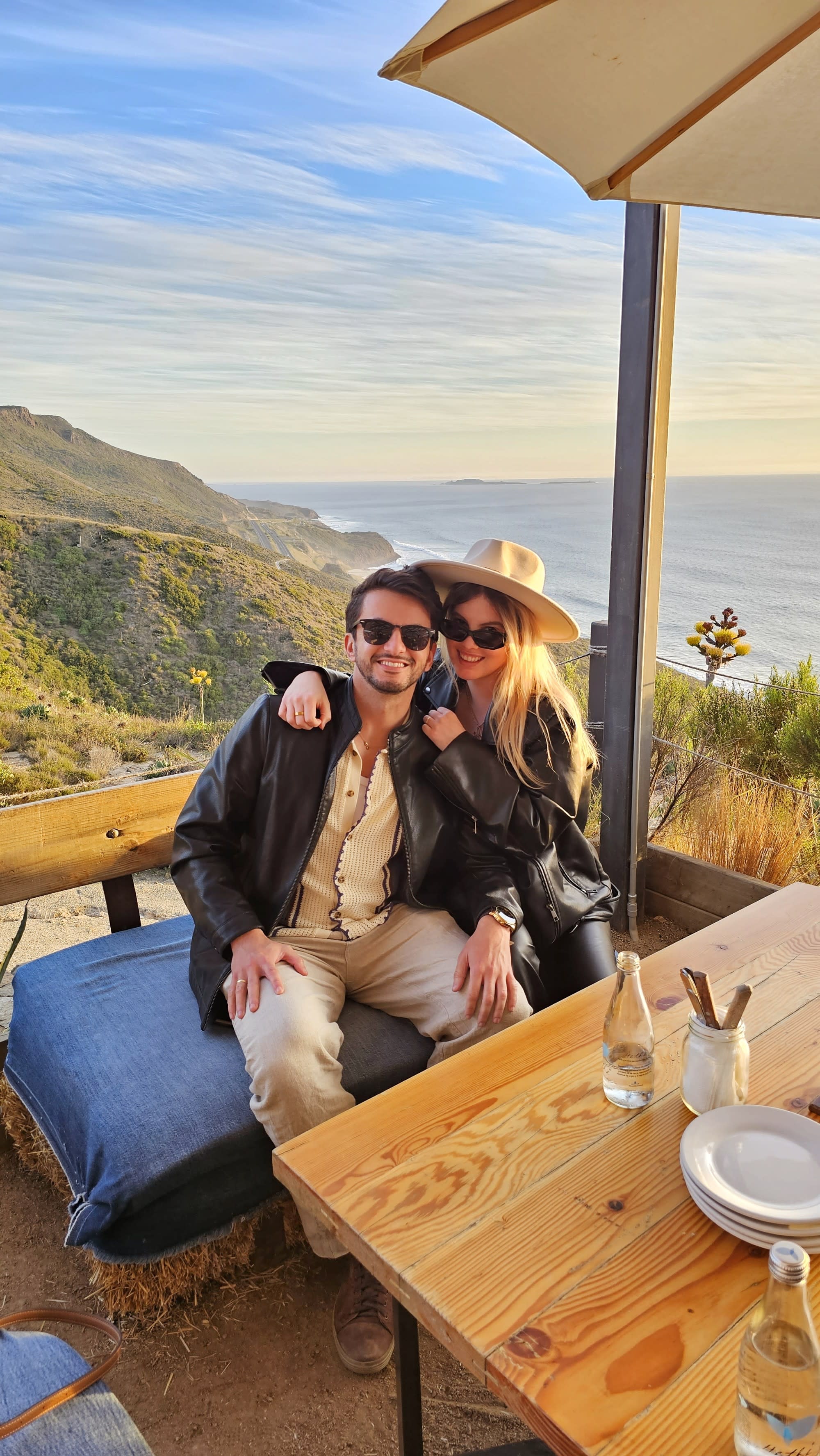 The image portrays two individuals seated at a wooden table, enjoying a coastal sunset view, with privacy maintained by obscuring their faces.