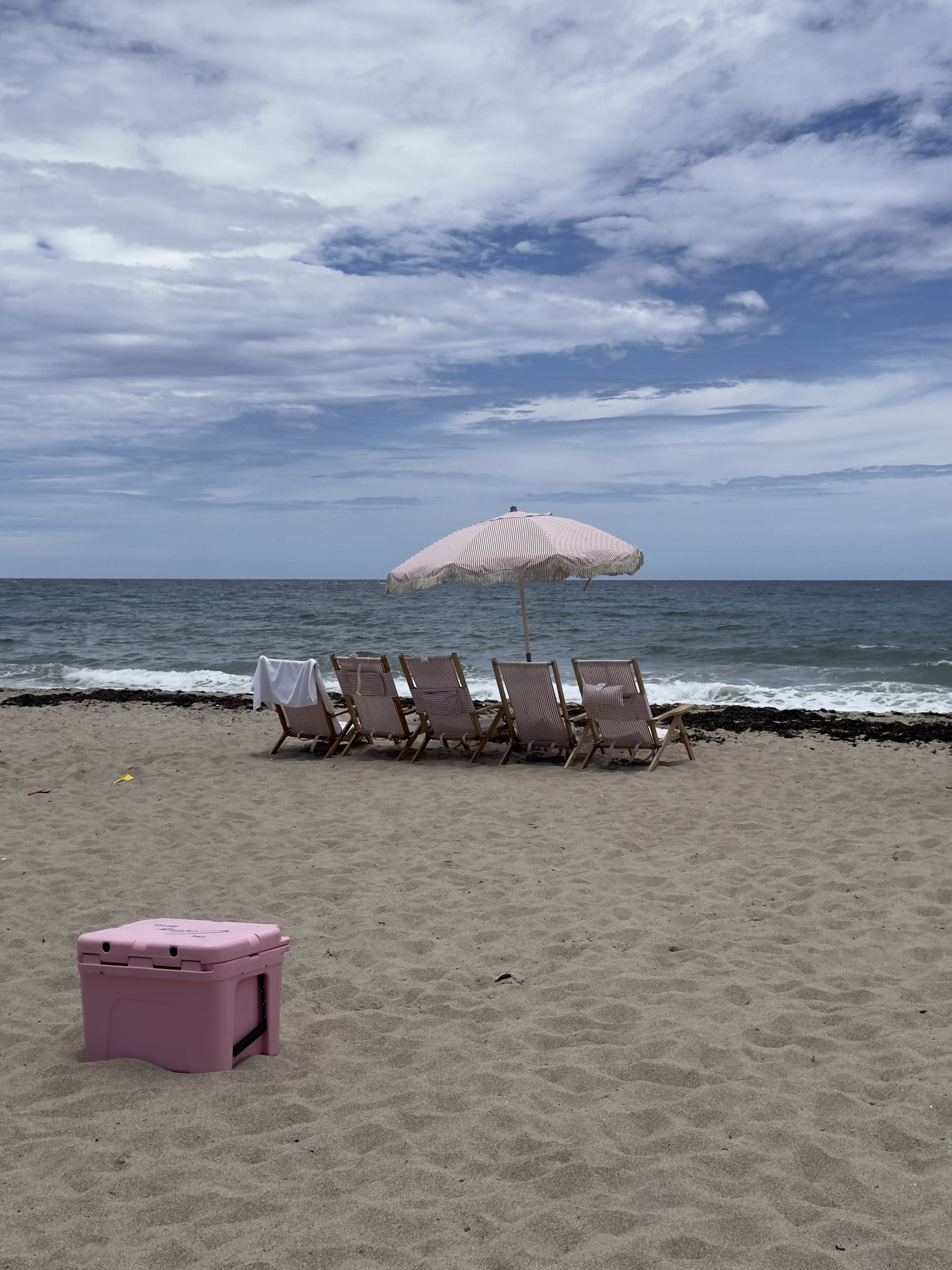 The image portrays a tranquil beach setting with a pink cooler, four wooden lounge chairs, and a striped sand umbrella overlooking the ocean.